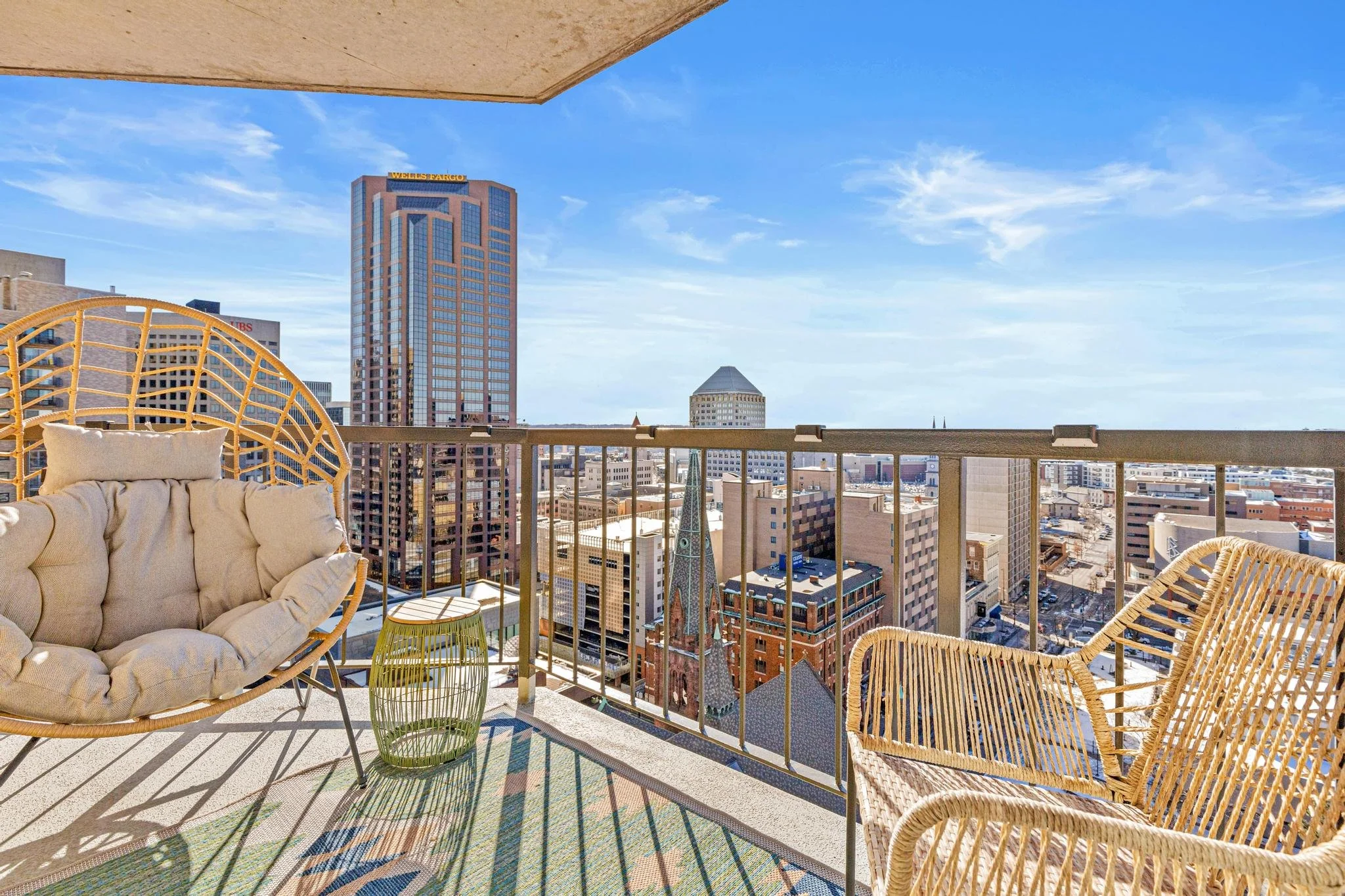 Balcony view overlooking city skyline with tall buildings, blue sky, and wispy clouds, featuring wicker chairs, a cushioned outdoor sofa, and a small green side table.
