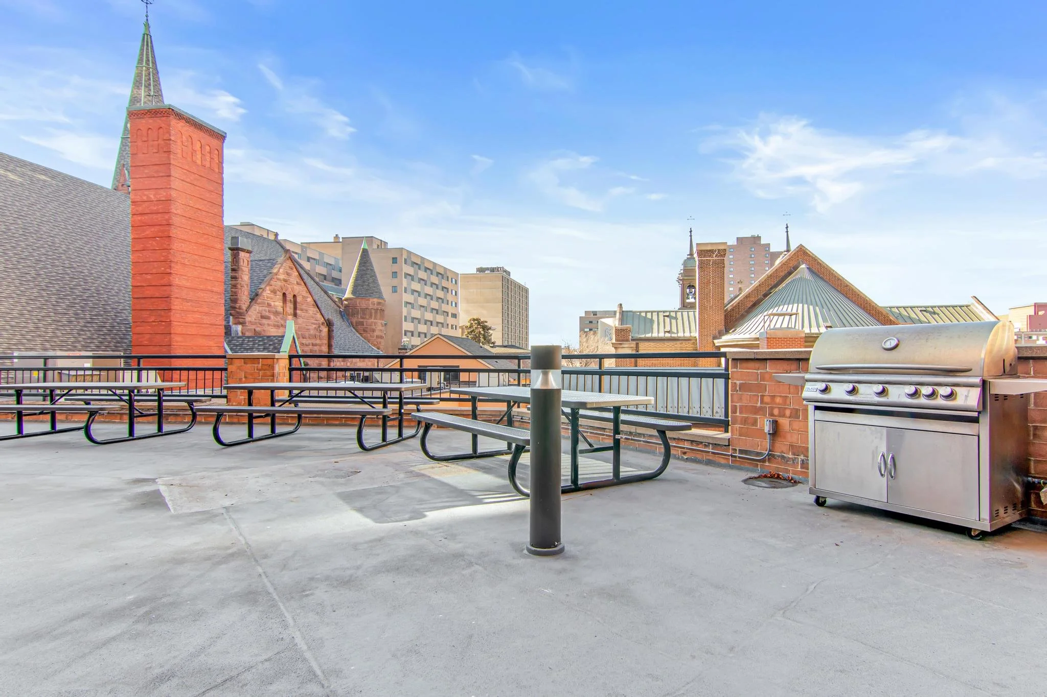 Rooftop with picnic tables, a grill, and a cityscape background with buildings and a church steeple under a blue cloudy sky.