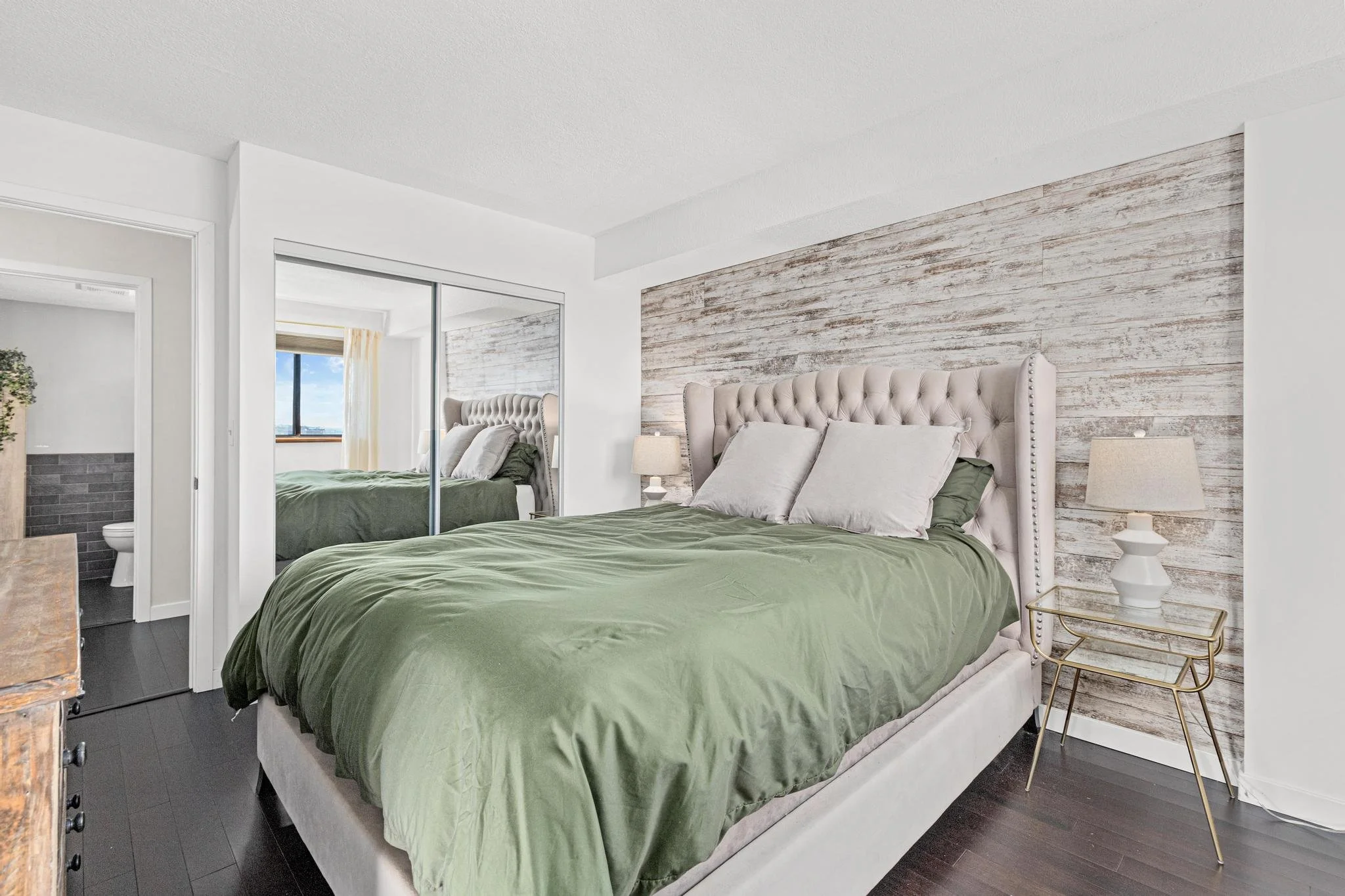 Modern bedroom with beige upholstered bed, green bedding, nightstand with lamp, mirrored closet, and view of additional bedroom area.