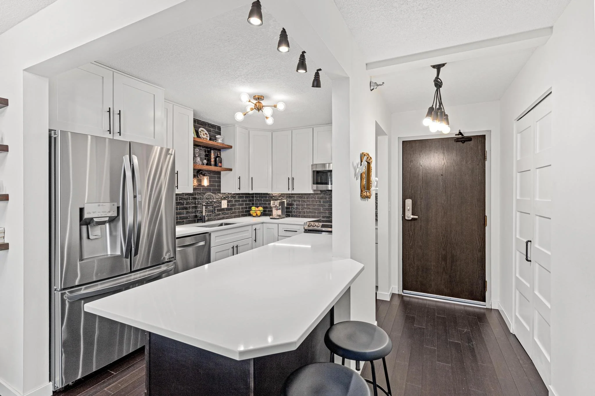 Modern kitchen with white cabinets, stainless steel refrigerator, black backsplash, and dark hardwood floors an entry door with a dark wood finish, and a kitchen island with two black stools