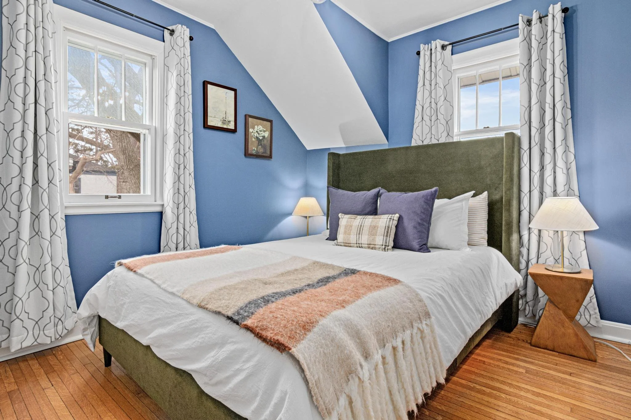 A cozy bedroom with blue walls, a green upholstered headboard, and hardwood floors. The bed has white sheets, purple and beige throw pillows, and a plaid lumbar pillow. Two table lamps are on either side of the bed, and two windows with white pattern