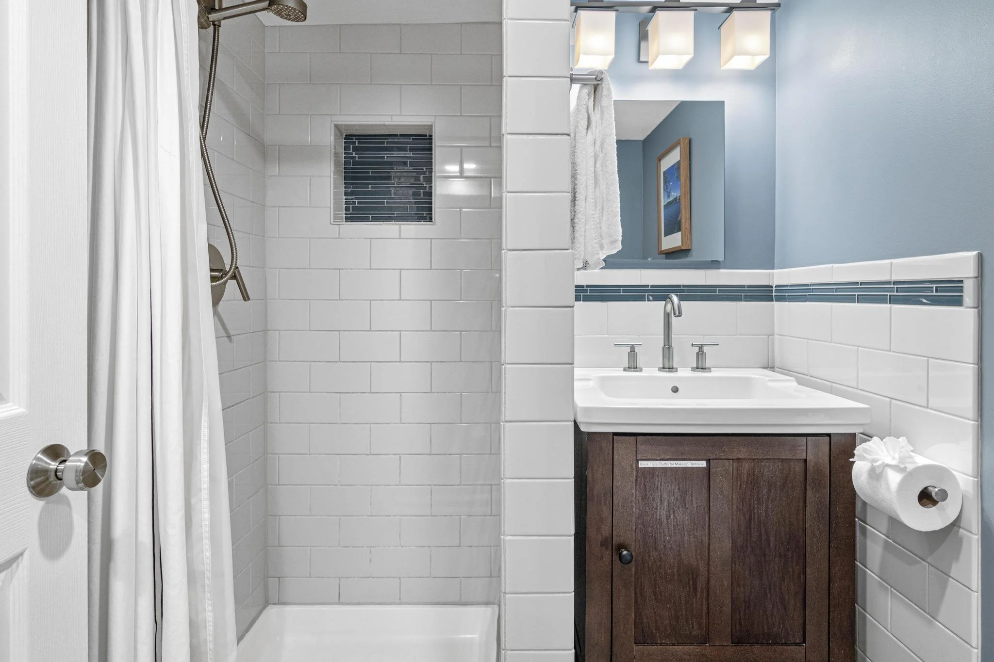 Bathroom with a shower to the left and a sink with a wooden cabinet below, a mirror with lighting above, and a blue wall with framed picture to the right.