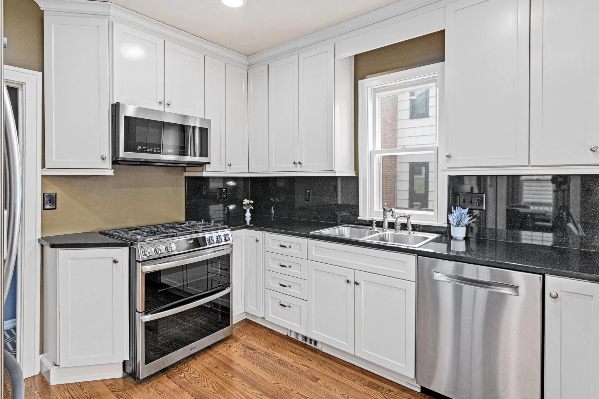 A modern kitchen with white cabinets, black countertops, a stainless steel oven and dishwasher, a microwave, and a double sink beneath a window.