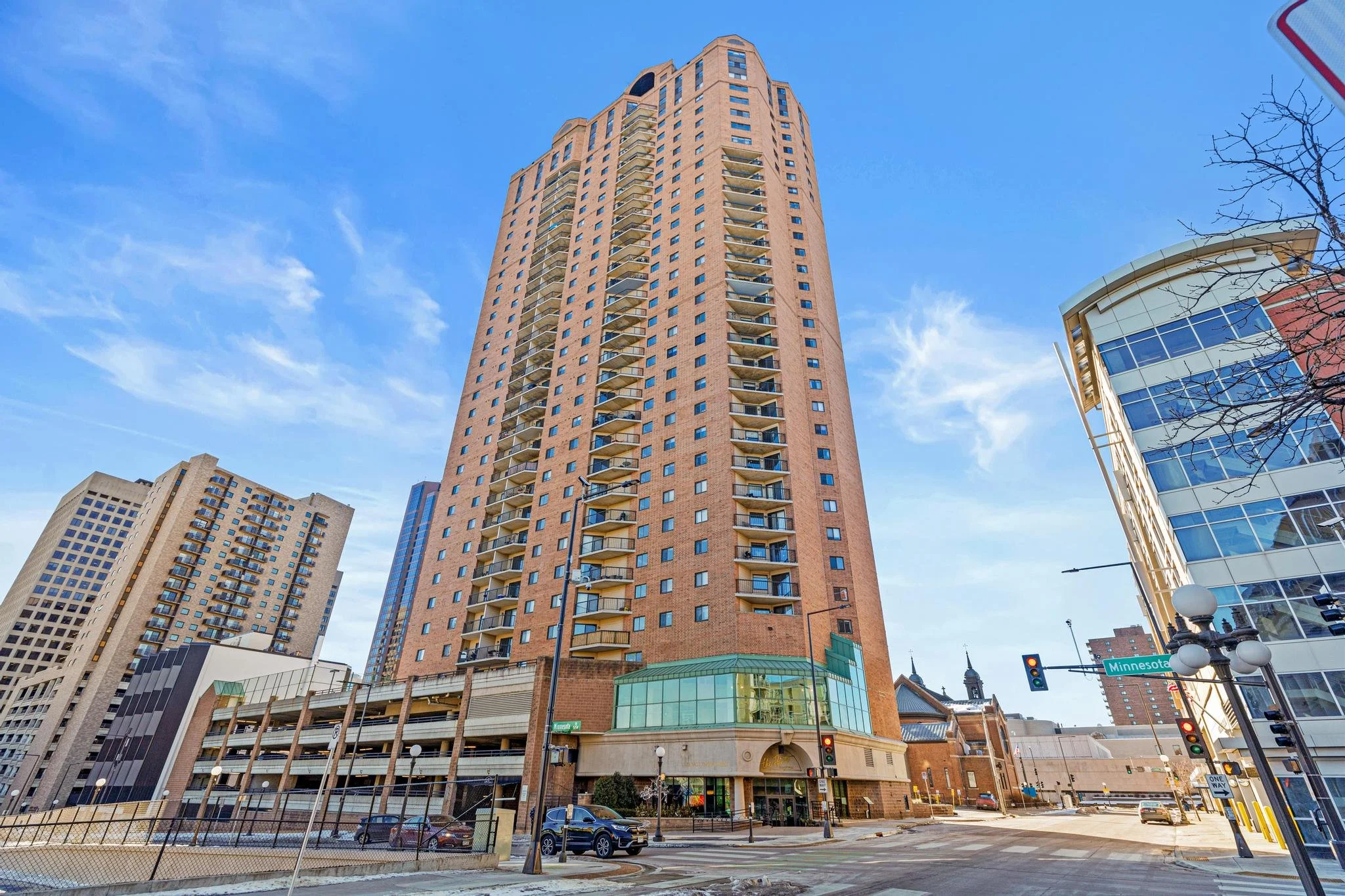 Tall brick residential building with balconies, surrounded by other high-rise buildings, in an urban area with streets, cars, and traffic lights.