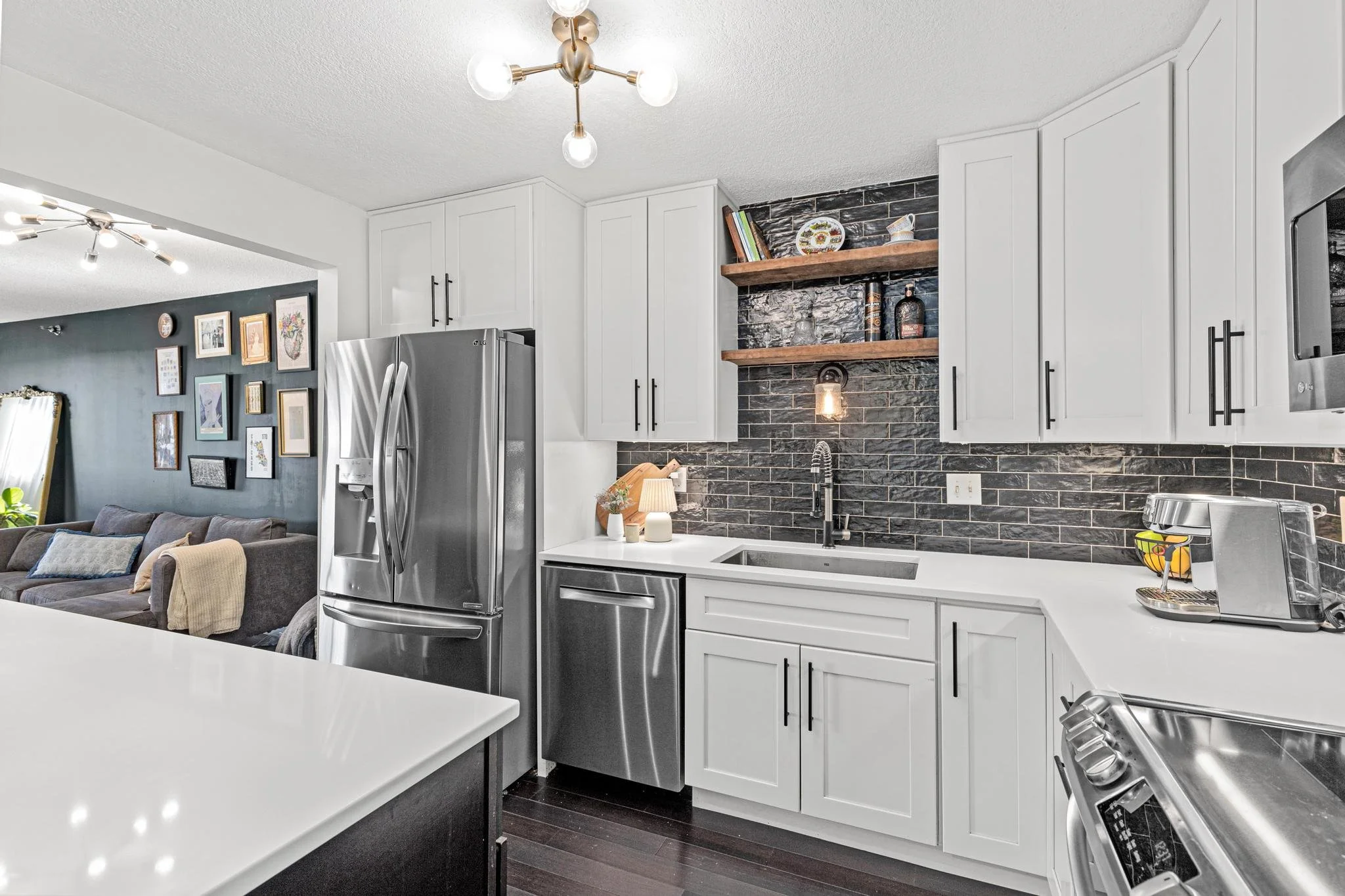 Modern kitchen with white cabinets, stainless steel refrigerator, black brick backsplash, floating wooden shelves, and a white countertop. Open to a living room with framed art on dark green wall.