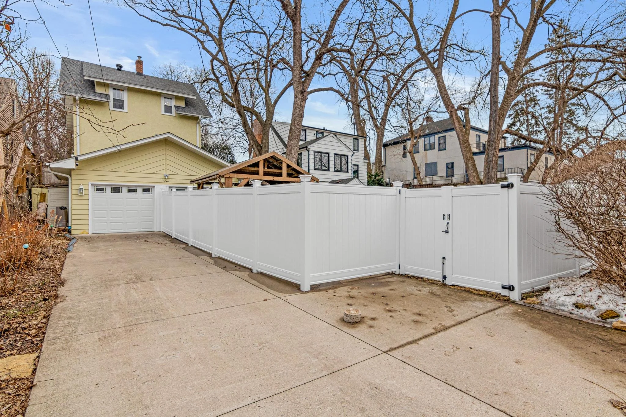 Backyard with a concrete driveway, white privacy fence, and trees without leaves; neighboring houses are visible in the background.