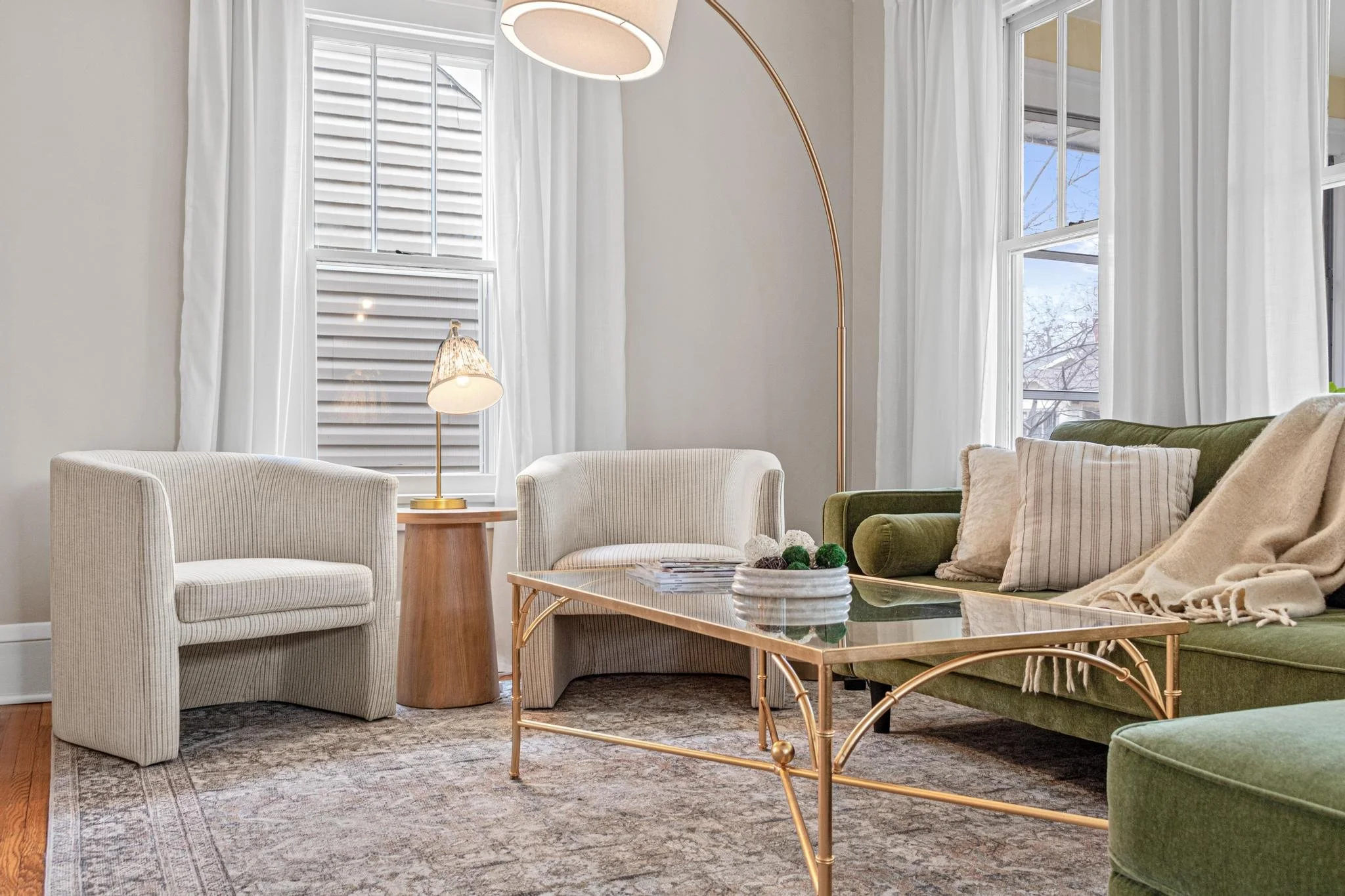 Living room with white and green furniture, gold coffee table, beige area rug, white curtains, and a tall white and gold floor lamp.