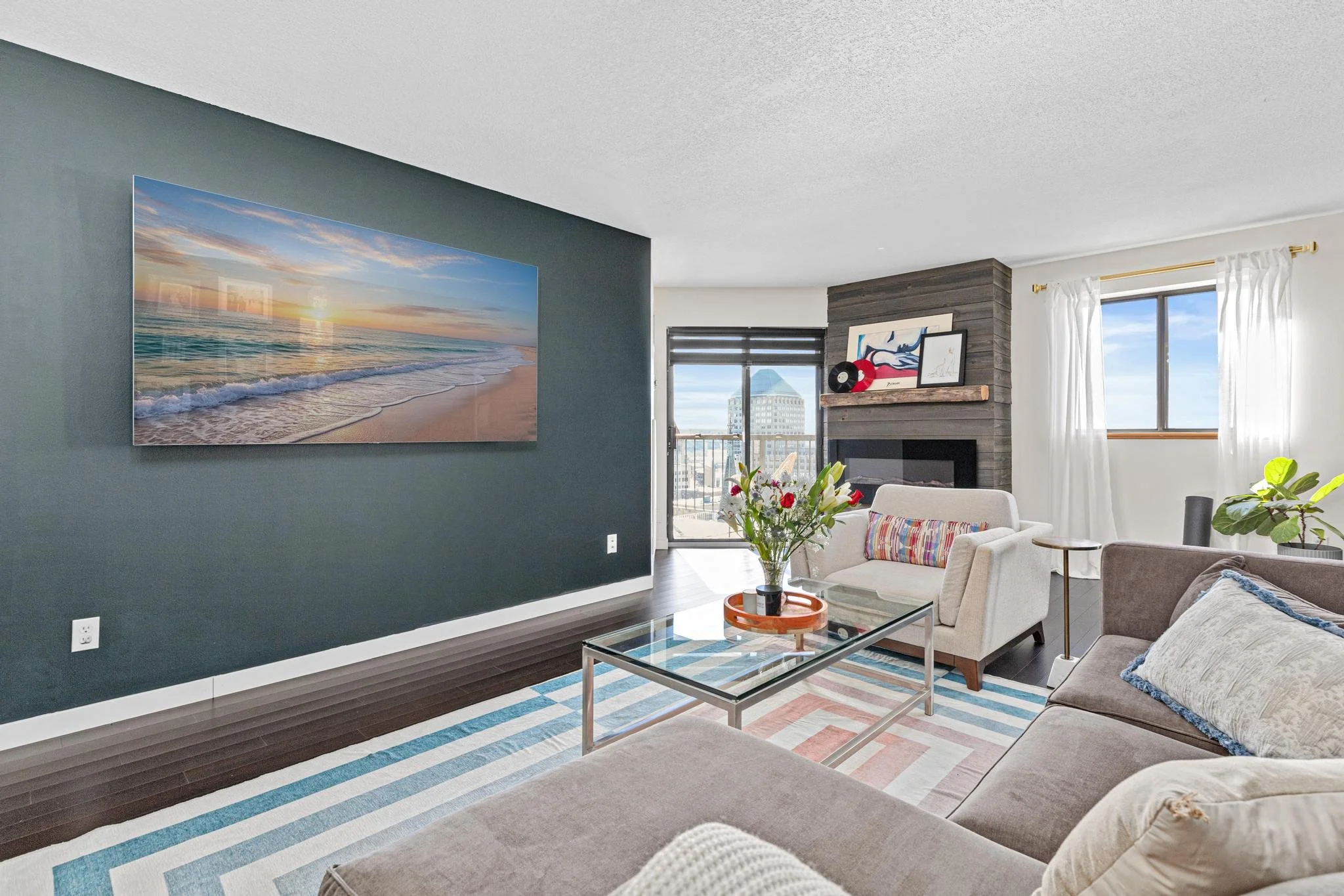 Living room with dark teal accent wall, large beach sunset picture, beige and gray sofas, glass coffee table with flower arrangement, fireplace with artwork, window with white curtains, balcony door with city view.