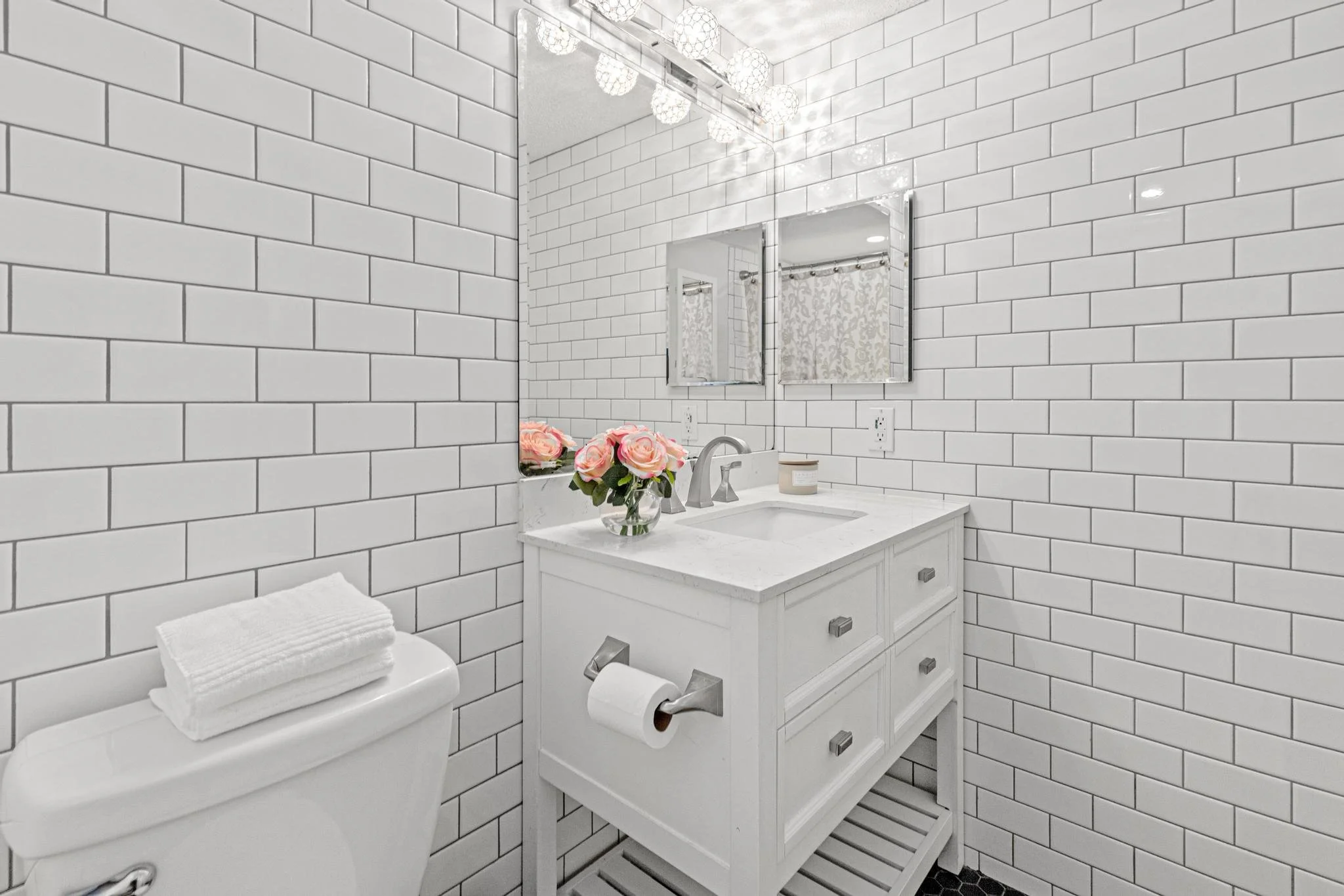 Small bathroom with white subway tile walls, white vanity with marble top, a rectangular mirror, a bouquet of pink roses, a framed mirror, a soap dispenser, and a toilet with a towel on top.