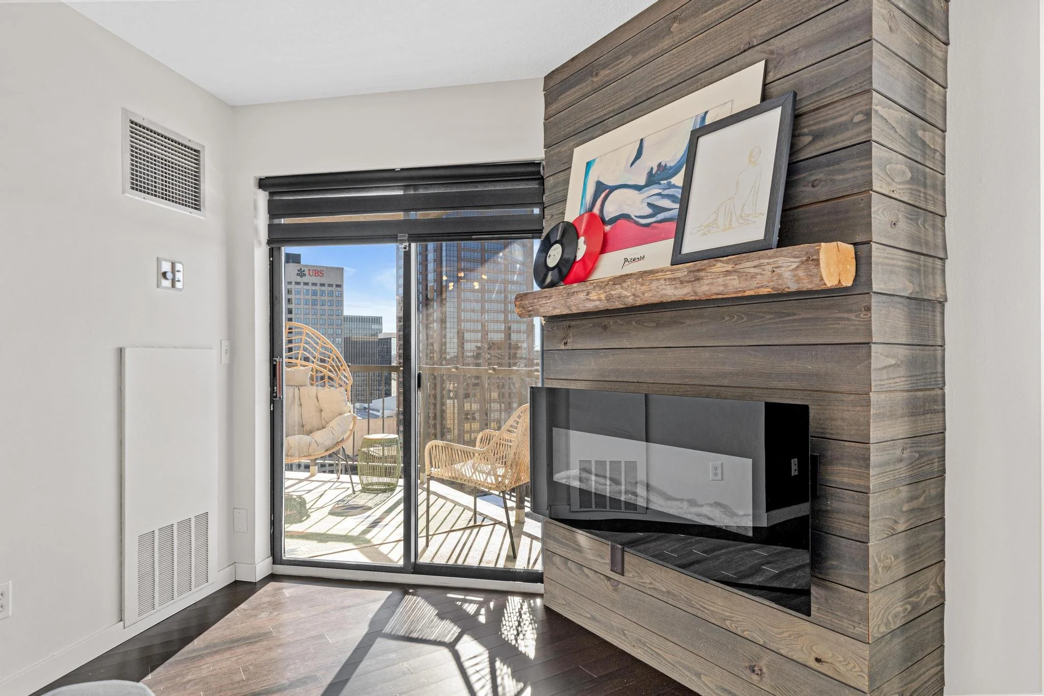 Living room with a wooden accent wall, fireplace, and art decorations above the mantel. Large sliding glass door leading to a balcony with outdoor seating and city view.