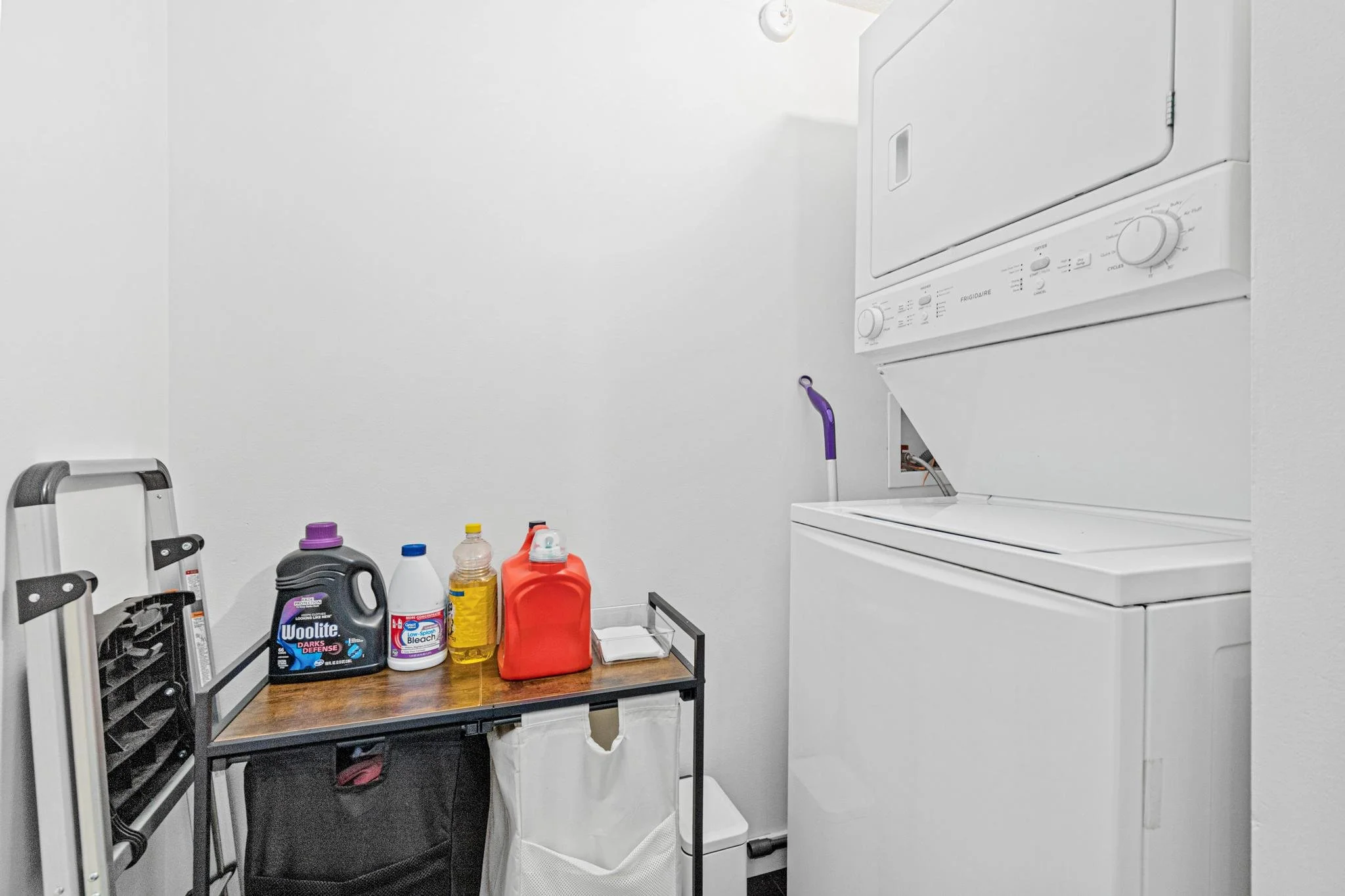 A laundry room with a stacked washer and dryer, laundry supplies on a small table, and a wall-mounted stepladder.