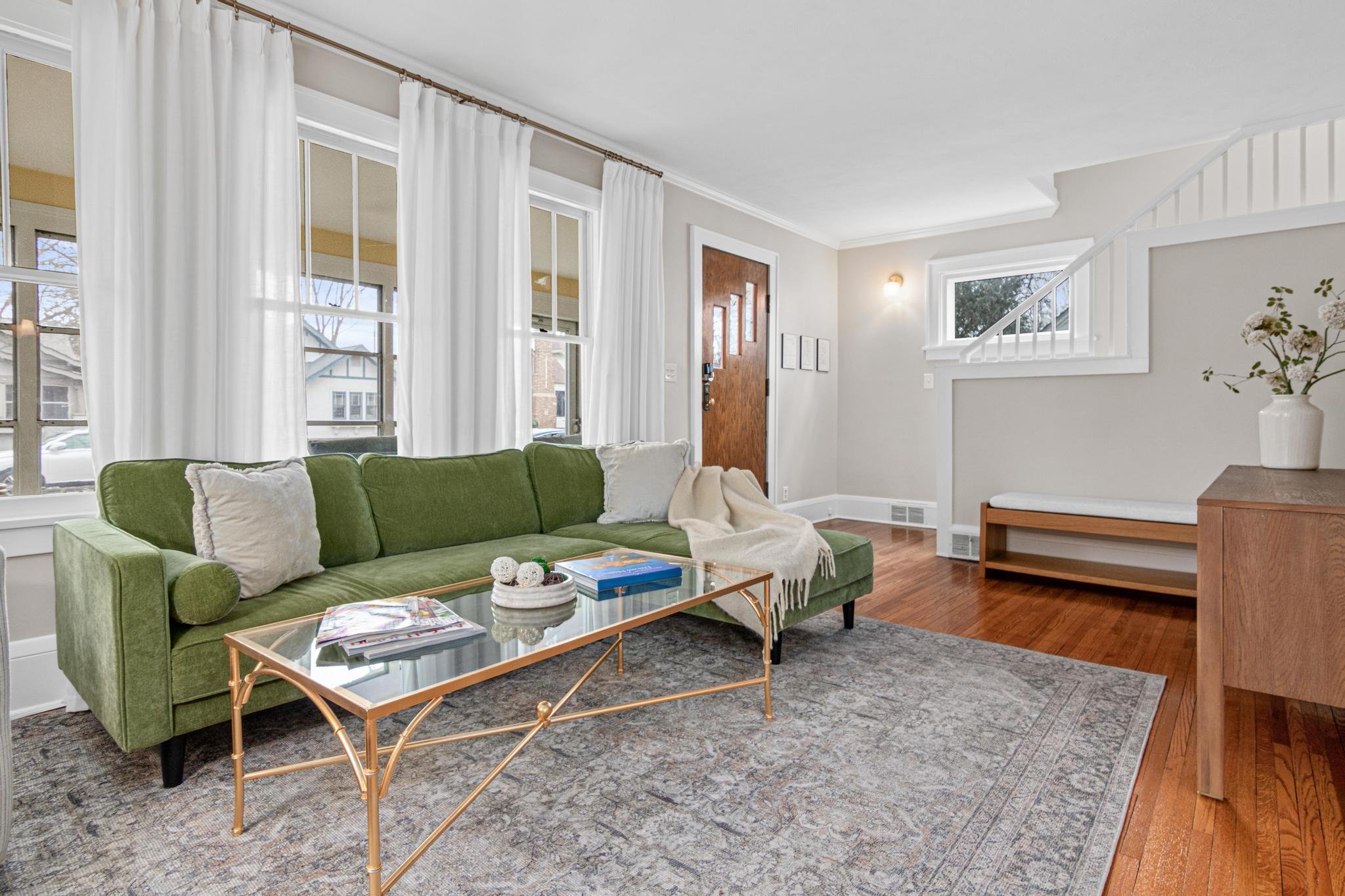 Living room with a green sofa, gold coffee table, white curtains, wooden door, wood flooring, and a decorative vase with flowers.