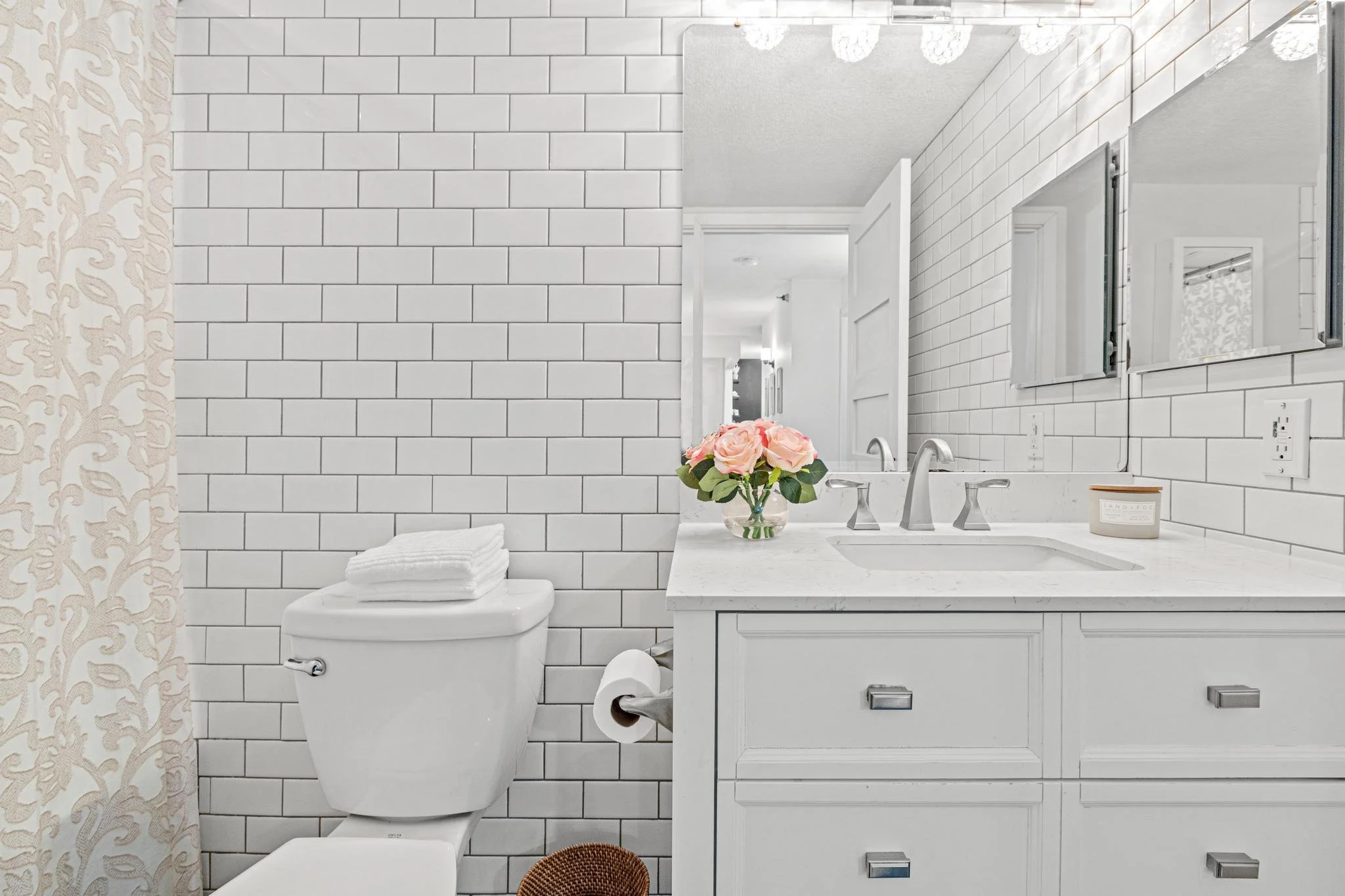 A modern bathroom with white subway tile walls, a white toilet with folded towels on top, a white vanity with a marble countertop, a vase of pink roses, and a mirror above the sink.