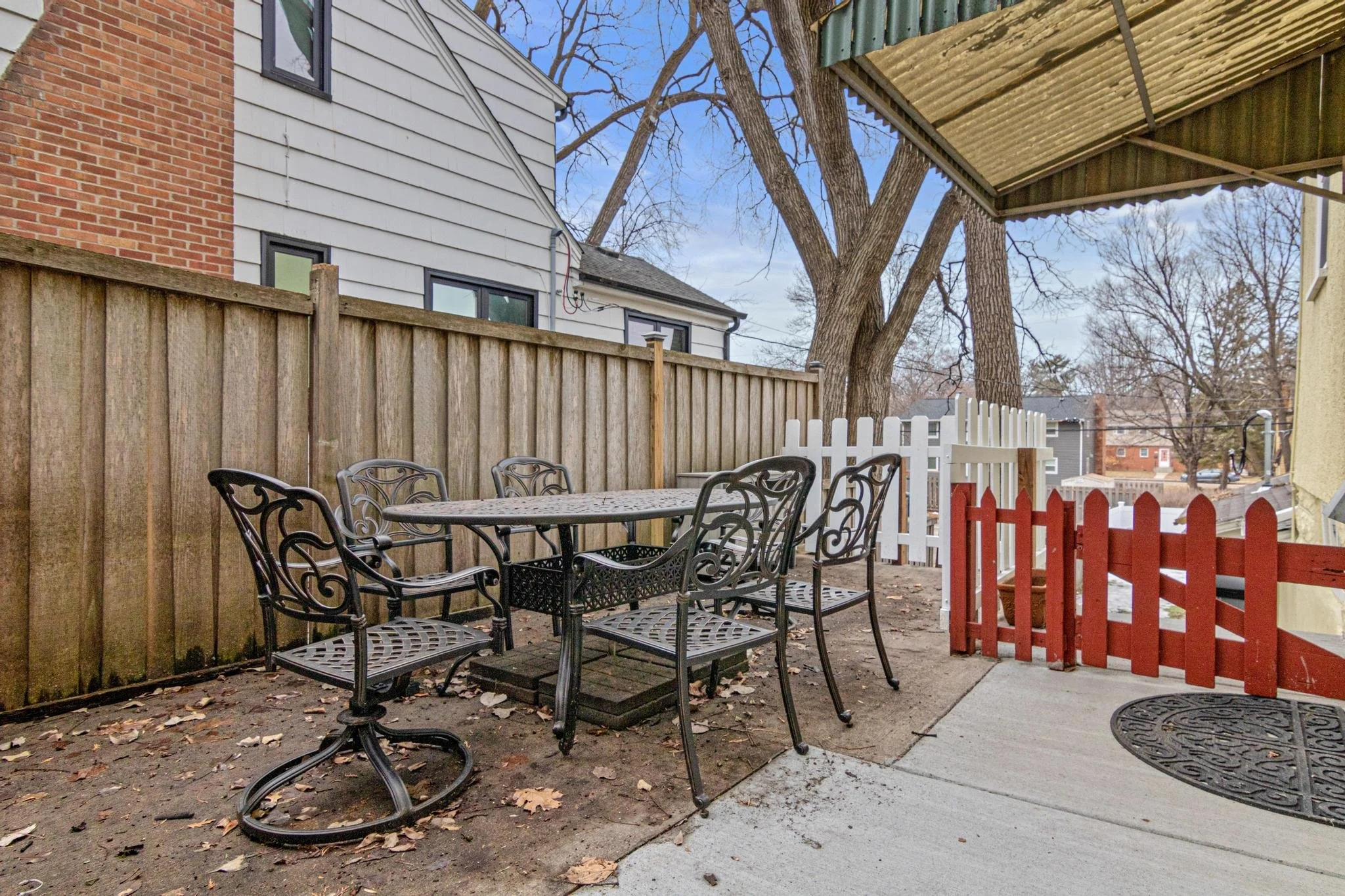 Backyard patio with black metal table and six chairs, surrounded by a wooden fence, with leaf-covered dirt ground and a concrete walkway. A white picket fence and a large tree are in the background, along with neighboring houses and trees.