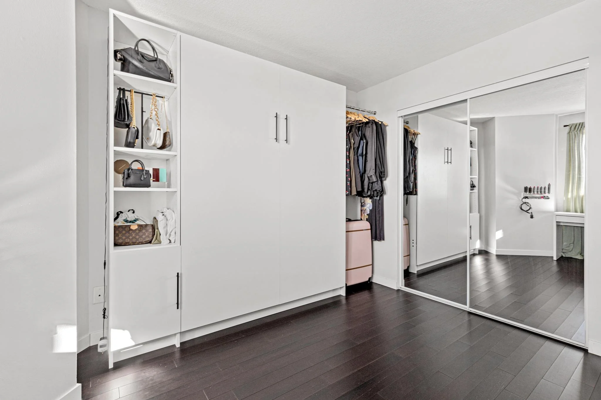 A walk-in closet with white cabinetry, a mirrored sliding door, hanging clothes, and shelves with handbags and accessories on a dark wood floor.