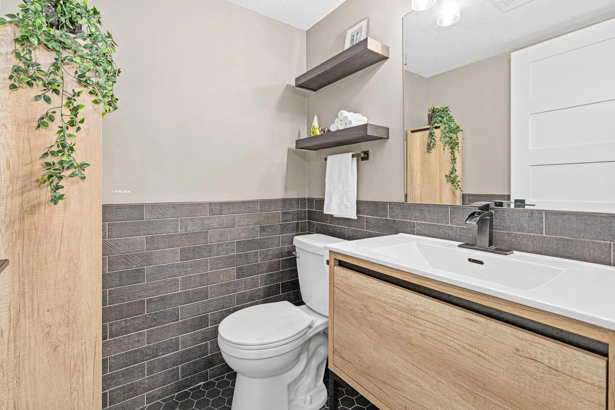 Modern bathroom with a toilet, wooden vanity, large mirror, black faucet, floating shelves with towels and decor, plant, and neutral colors.