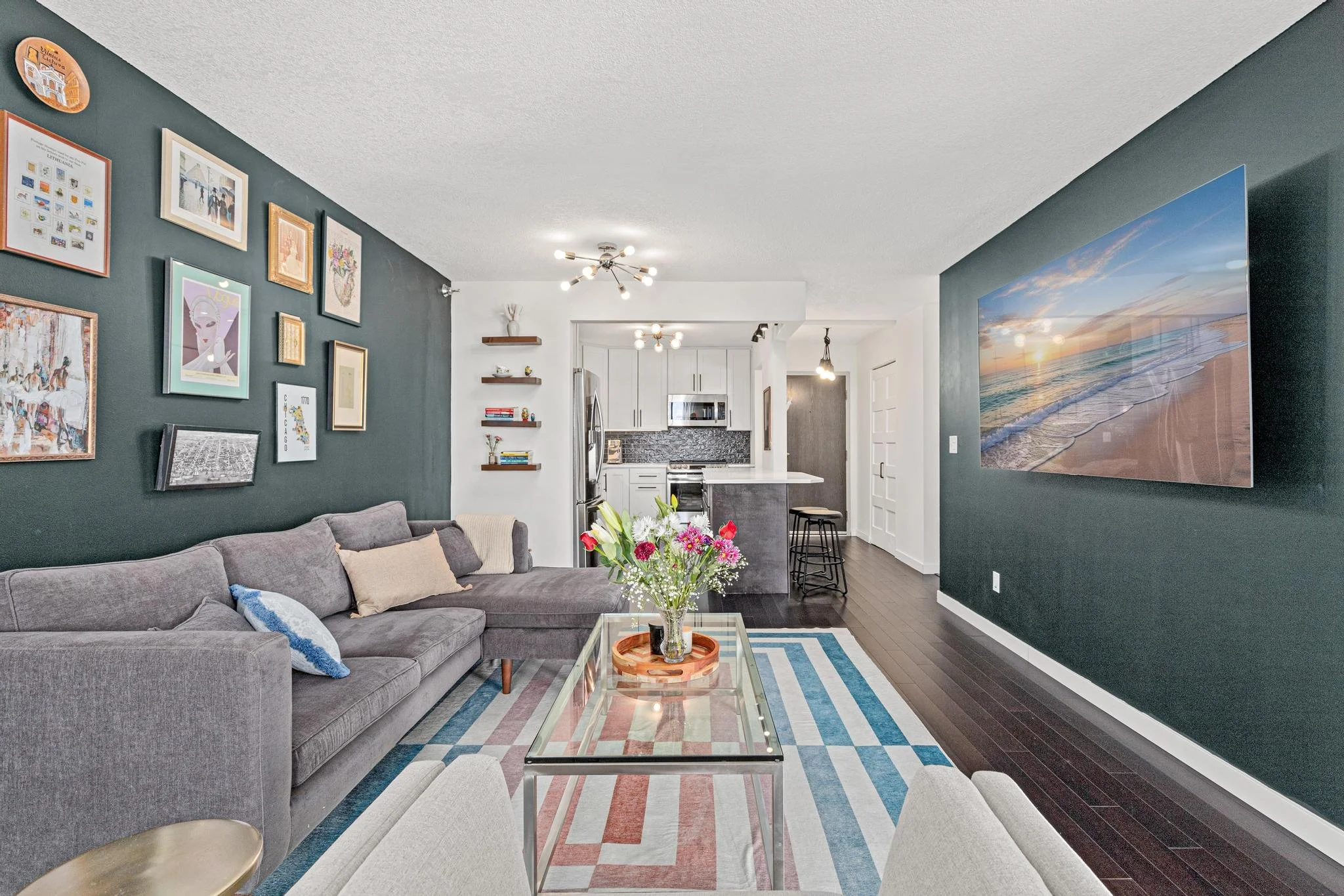Modern living room with dark green accent walls, artwork, a gray sectional sofa with cushions, a glass coffee table with a flower arrangement, and a large wall-mounted flat-screen TV displaying a beach sunset. The open kitchen area has white cabinets