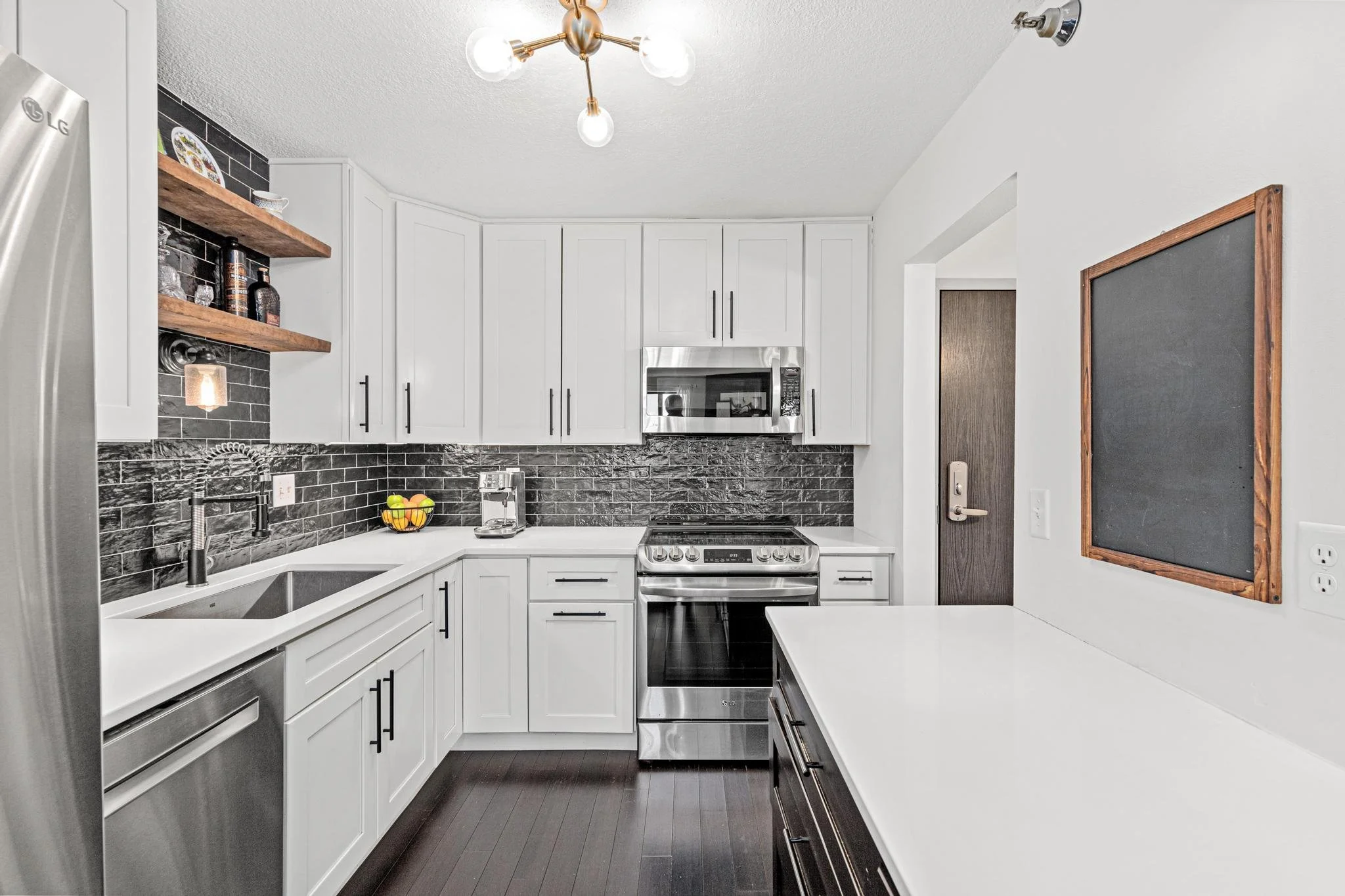 Modern kitchen with white cabinets, black backsplash, stainless steel appliances, open shelving, dark hardwood floors, and a small blackboard on the wall.