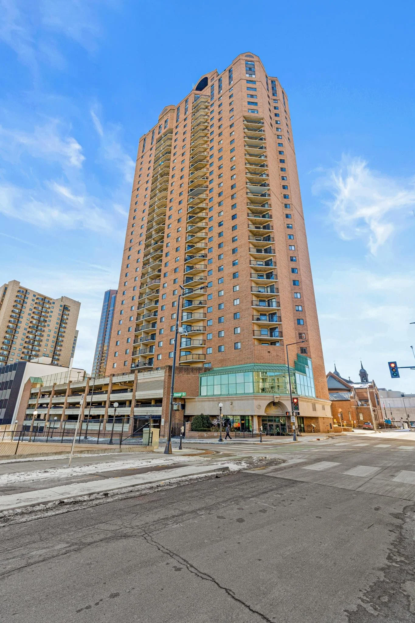 A tall brick residential building with balconies on each floor, situated at an urban street corner with traffic lights, crosswalks, and other high-rise buildings in the background under a blue sky.