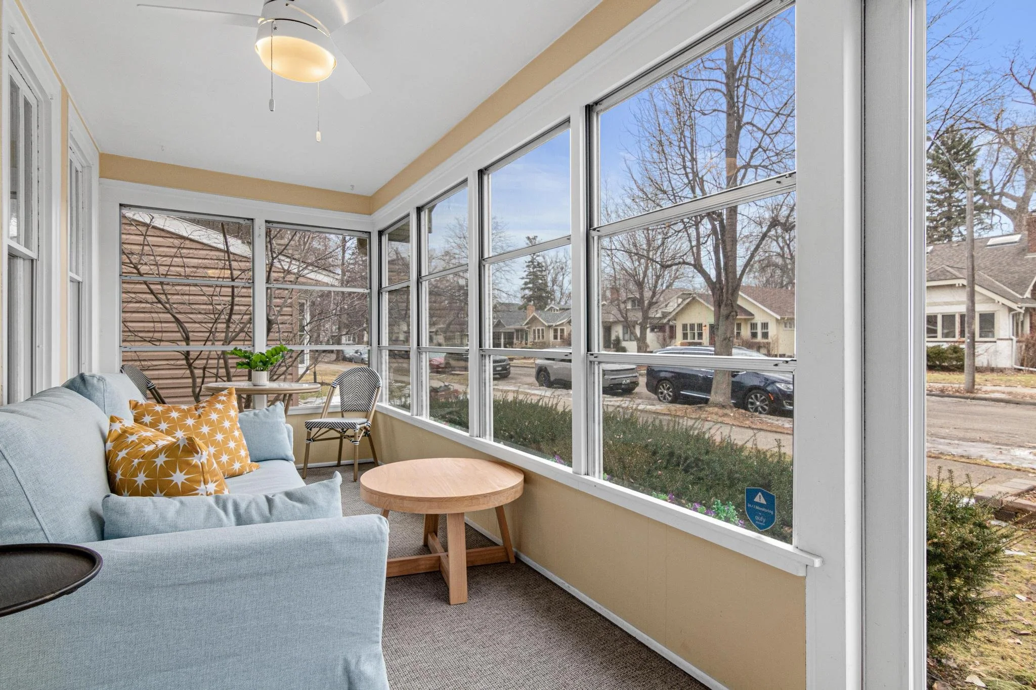 Sunroom with blue sofa, yellow star-patterned pillows, small round wooden table, potted plant, and large windows showing a suburban street scene with trees and houses.