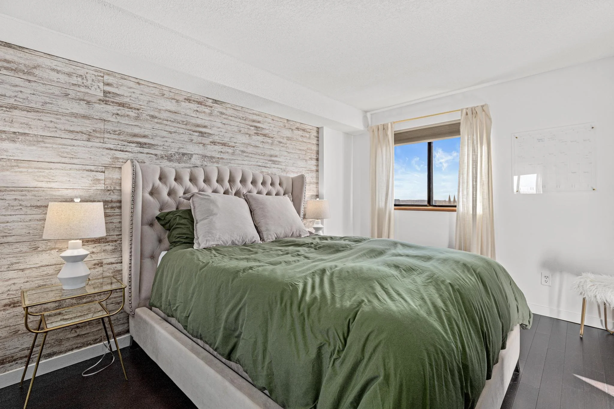 A bedroom with a large bed covered in a green blanket, gray pillows, a tufted headboard, two bedside lamps, a window with cream curtains, and a white wall with a whiteboard and a small white furry stool.