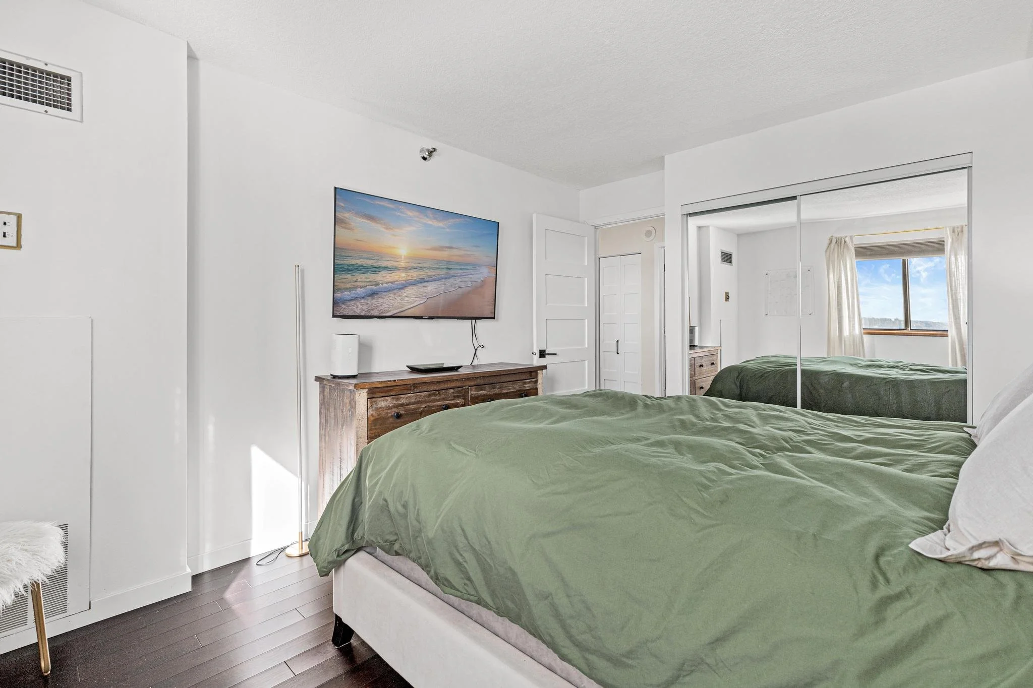 Bedroom with a bed covered in green bedding, a mirrored closet, a wooden dresser beneath a wall-mounted TV showing a beach scene, and a window with curtains showing a blue sky and clouds.