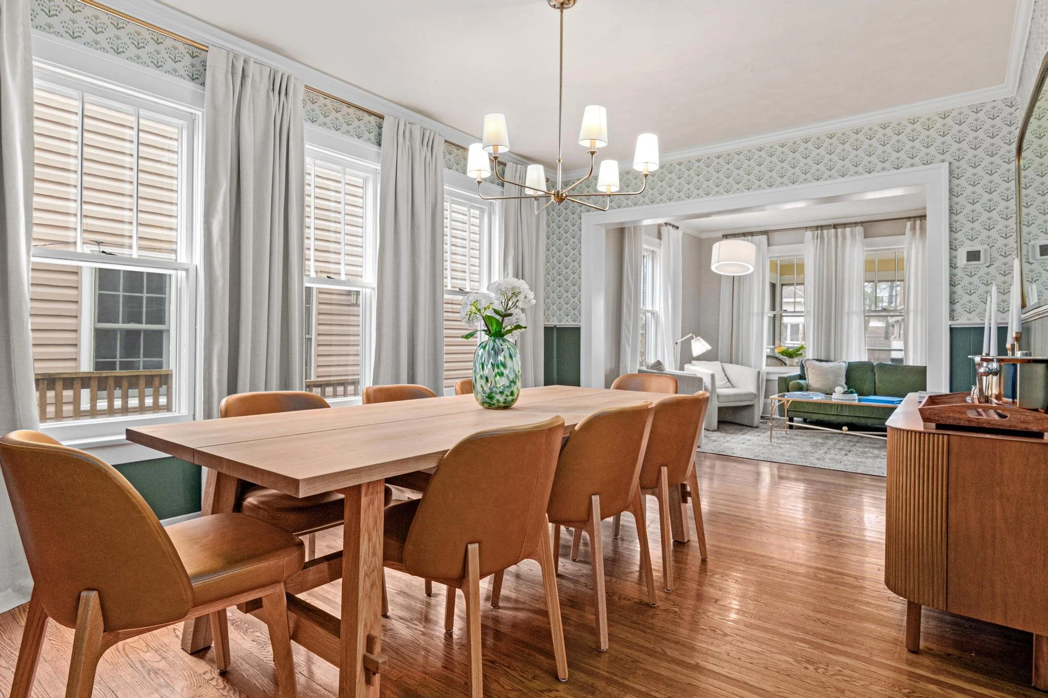 A dining room with a wooden table and brown chairs, a vase with white flowers on the table, large windows with white curtains, and a chandelier hanging from the ceiling. In the background, there is a living room with white sofas and a green sofa, a c