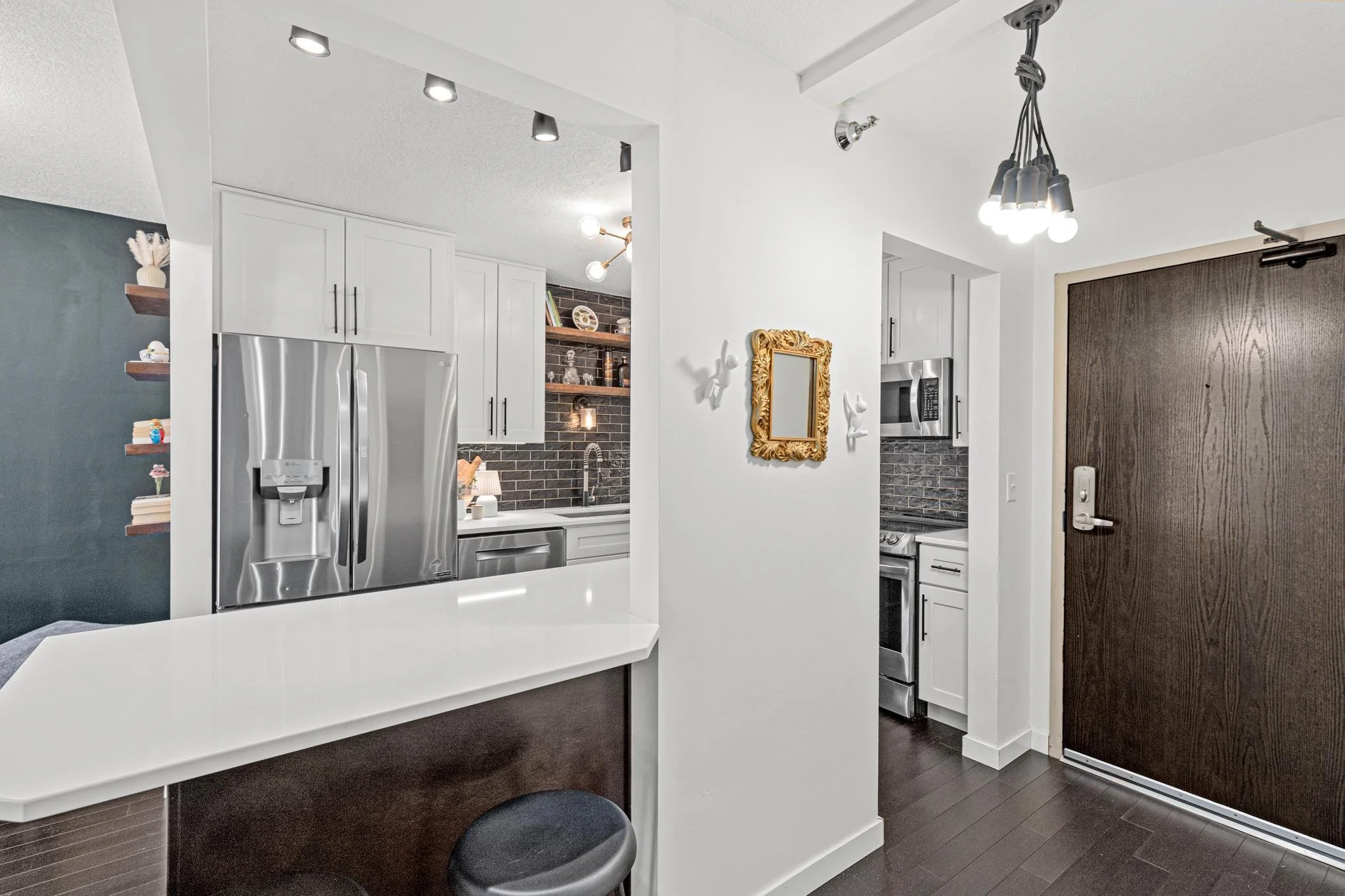 Modern kitchen with white cabinetry, stainless steel appliances, black brick backsplash, and dark wood floors. Part of a living area with dark accent wall and floating shelves.