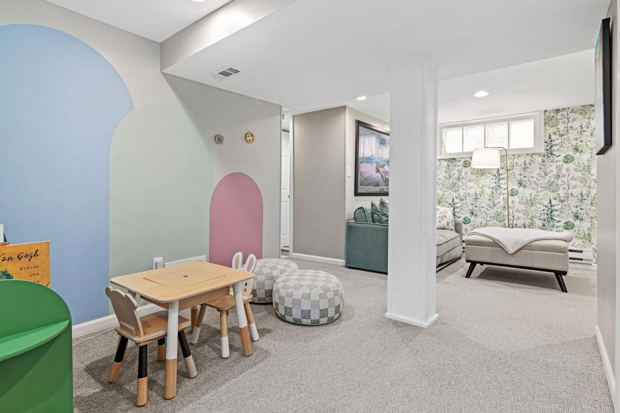 Children's play area with small table and chairs, patterned poufs, and a colorful wall mural, adjacent to a cozy living room with a patterned accent wall and seating.