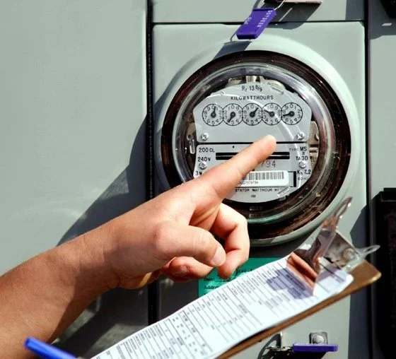 Close-up of a person pointing at an electric submeter with a clipboard in hand outside of a building.