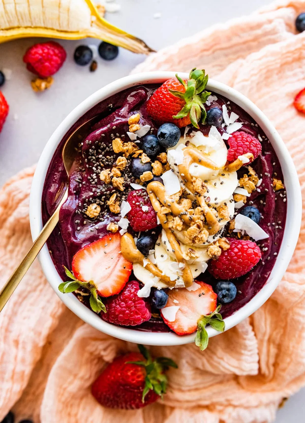 A bowl of smoothie or yogurt topped with strawberries, raspberries, blueberries, granola, chia seeds, coconut flakes, and a drizzle of peanut butter, on a light surface with peach-colored cloth and additional berries around.