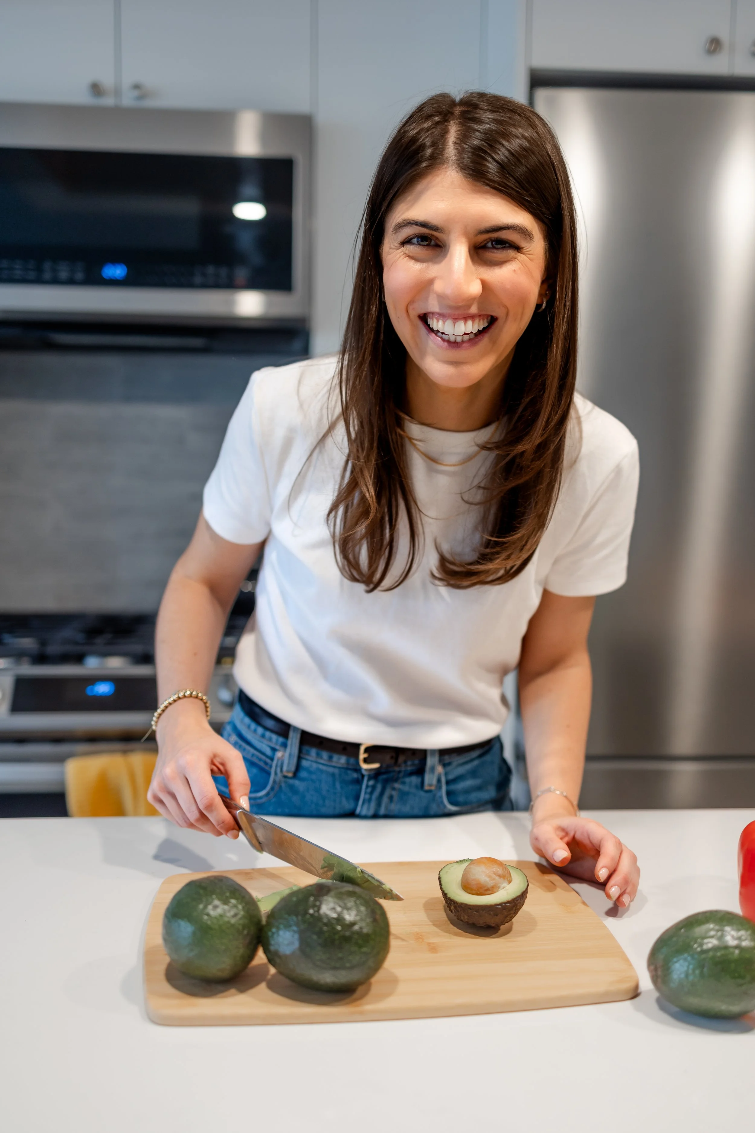 A woman cutting an avocado in a kitchen, smiling at the camera with a half avocado on the cutting board.