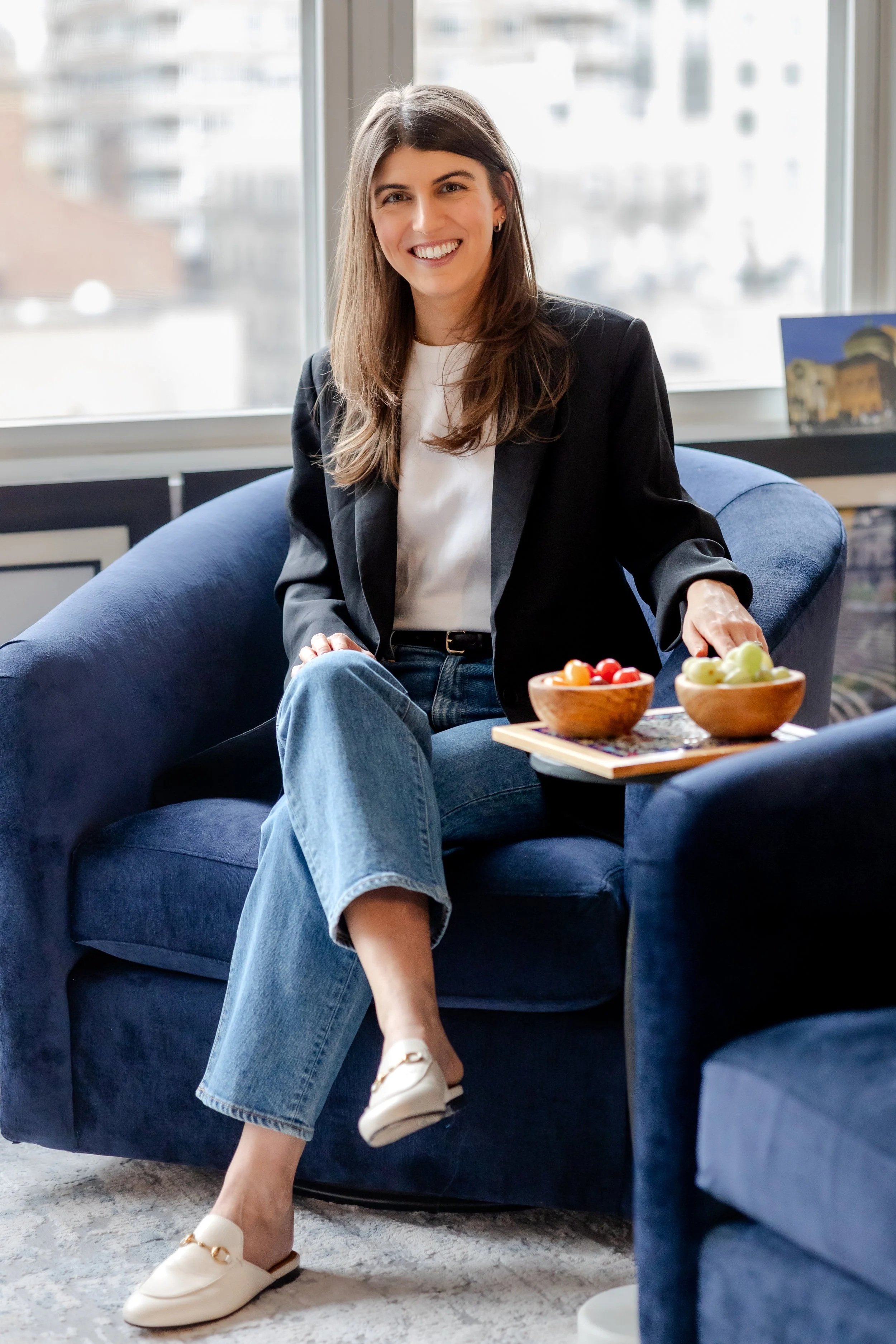 A woman with long brown hair, wearing a black blazer, white t-shirt, and jeans, sitting on a blue couch with a big window behind her, smiling, with bowls of fruit on a table beside her.