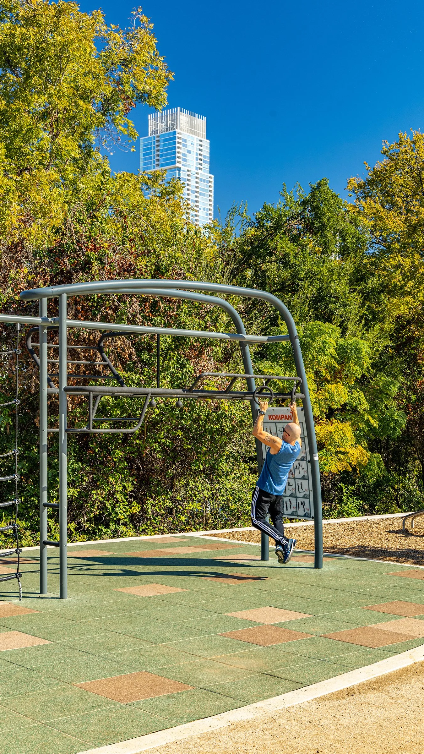 A man hanging from gymnastic rings at an outdoor playground with trees and a skyscraper in the background.