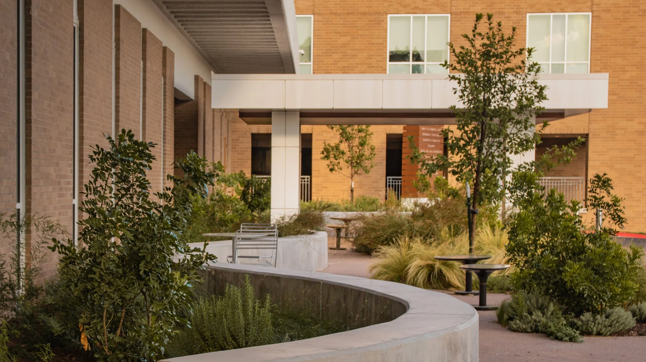 Outdoor courtyard with plants, trees, concrete benches, and a curved planter, surrounded by modern brick buildings and large windows.
