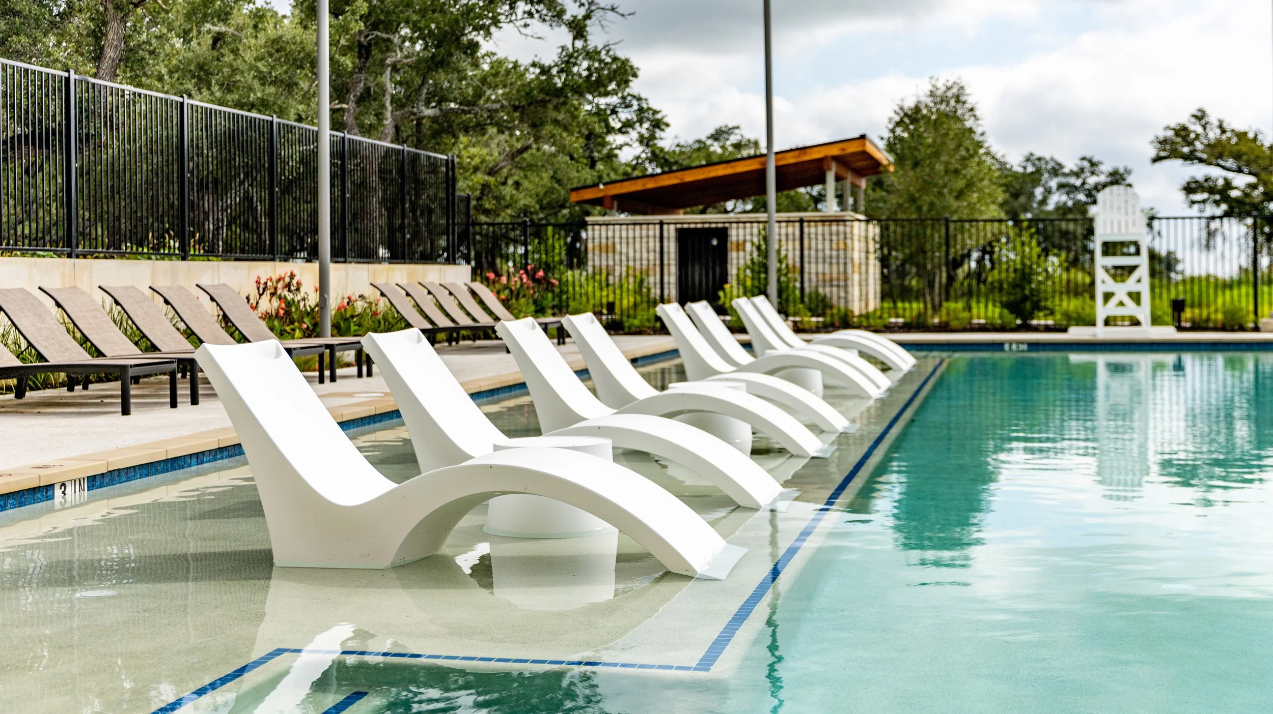 Empty swimming pool with white lounge chairs partially submerged in water, surrounded by a black metal fence, trees, and cloudy sky.