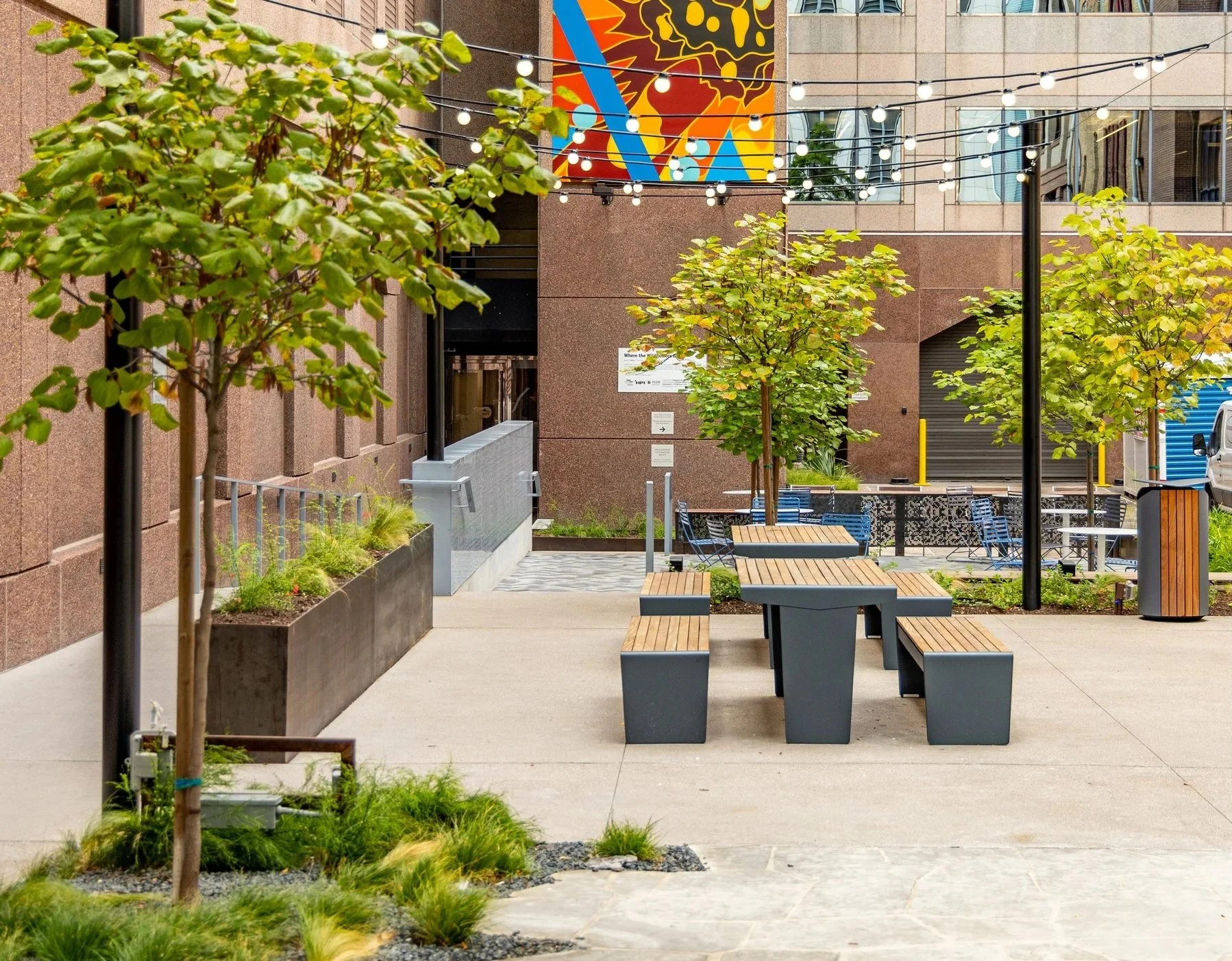 An outdoor urban area with young trees, benches, string lights, and a colorful mural on a building wall.