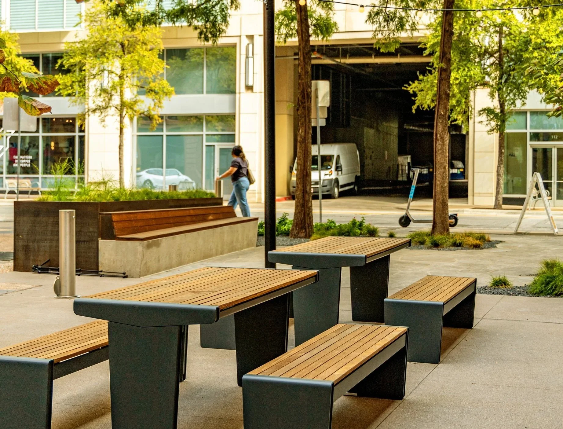 Outdoor urban scene with wooden benches and tables, trees, a person walking, a scooter, a parked van, and a building with large glass windows.