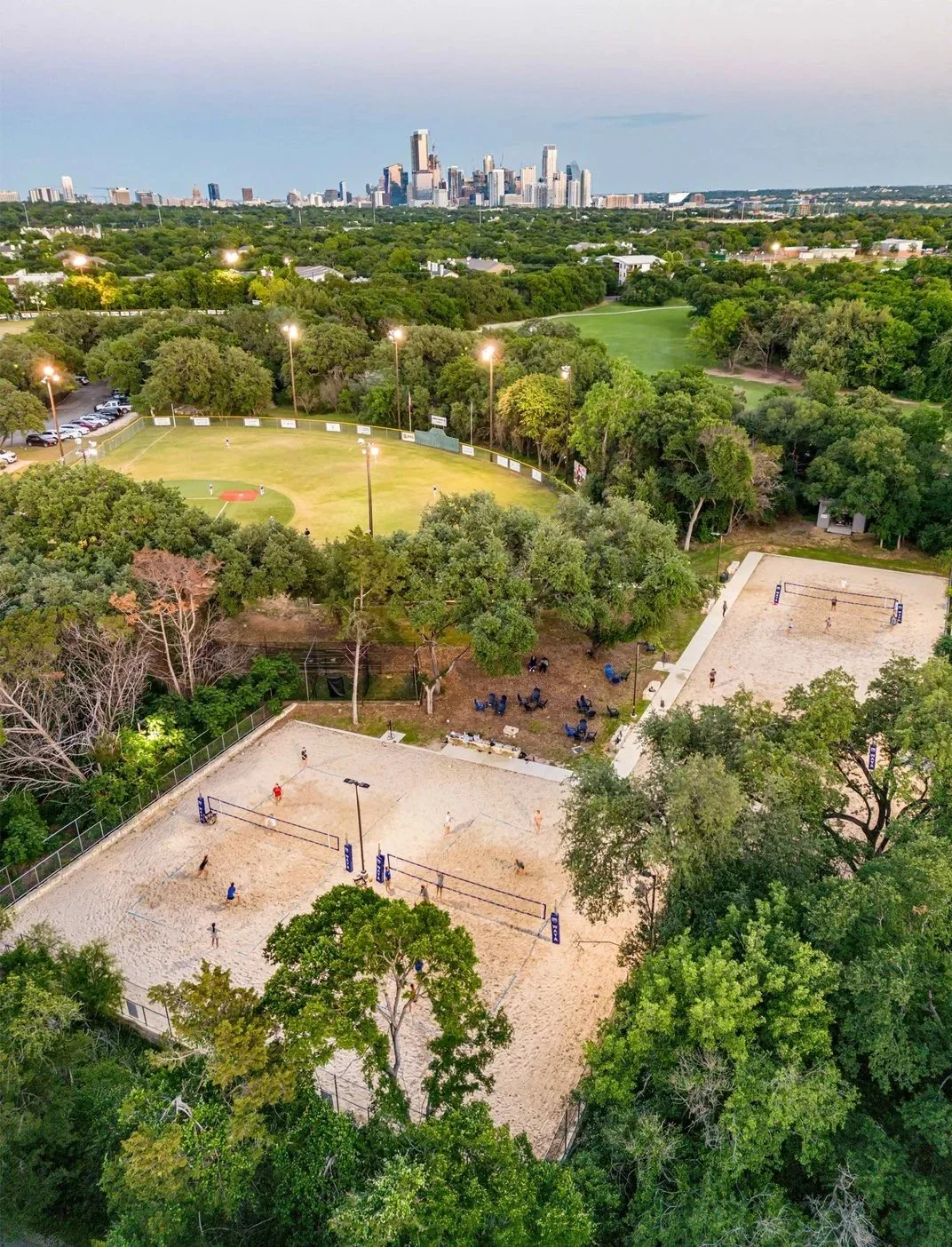 Aerial view of a sports complex with a baseball field, volleyball courts, and a soccer field, surrounded by trees and with a city skyline in the background.
