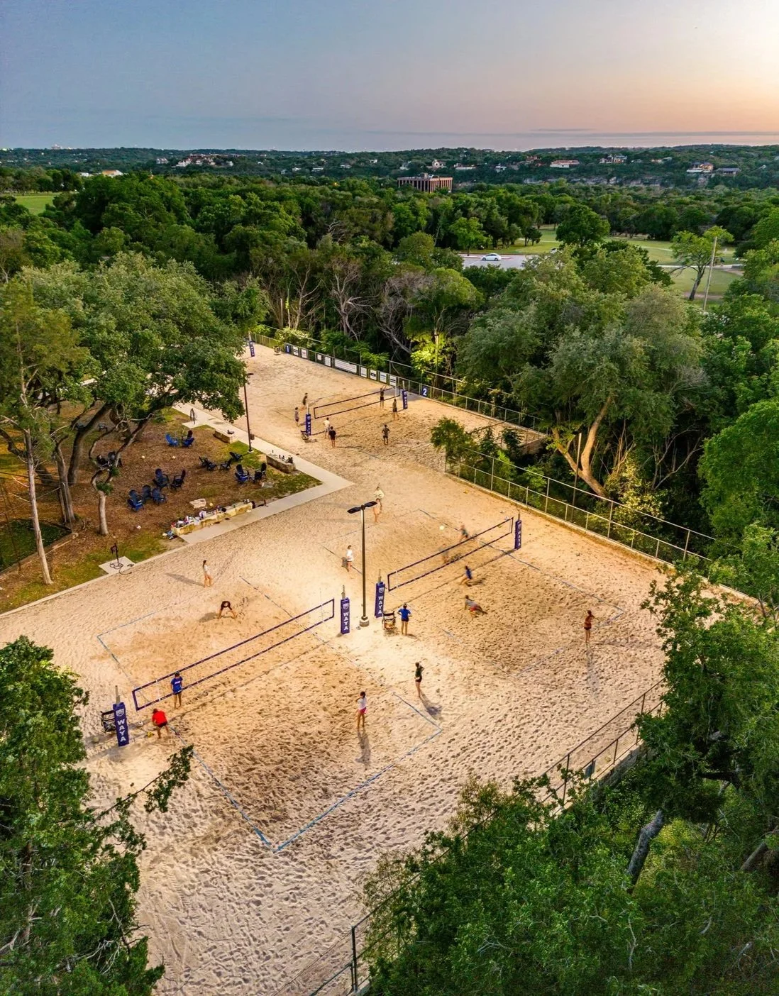 An aerial view of a sandy volleyball court with players playing, surrounded by trees and with a bench area nearby, during sunset with a distant treeline and sky.