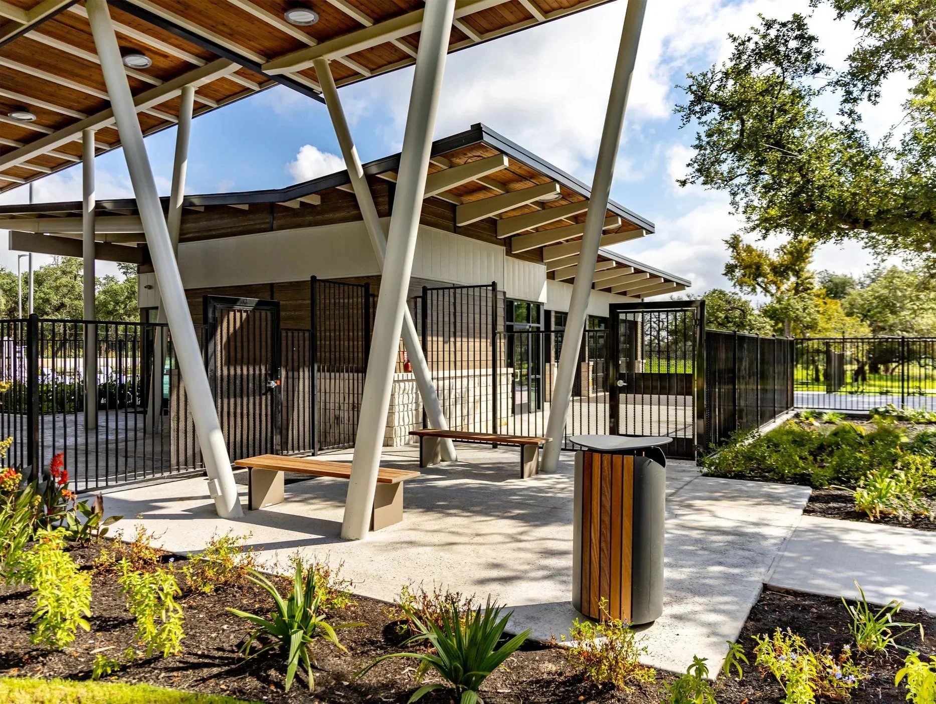Modern building with angled wooden roofs and black metal gates, surrounded by landscaped garden with flowers and trees, sunny day with partly cloudy sky.