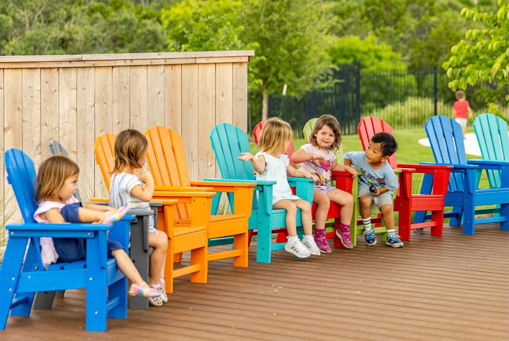Six children sitting on colorful Adirondack chairs on a wooden deck in a park with green trees and a grass field in the background.