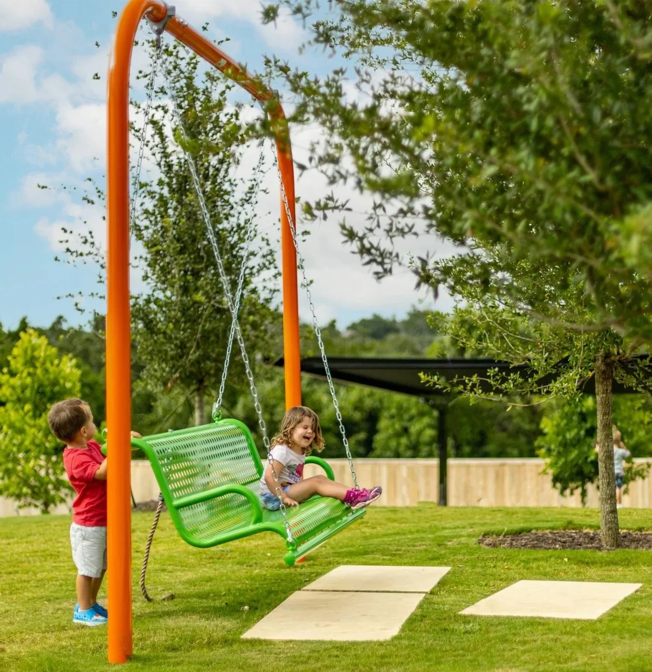 A boy pushes a green swing with a girl sitting on it in a park with trees and a fence, sunny weather.