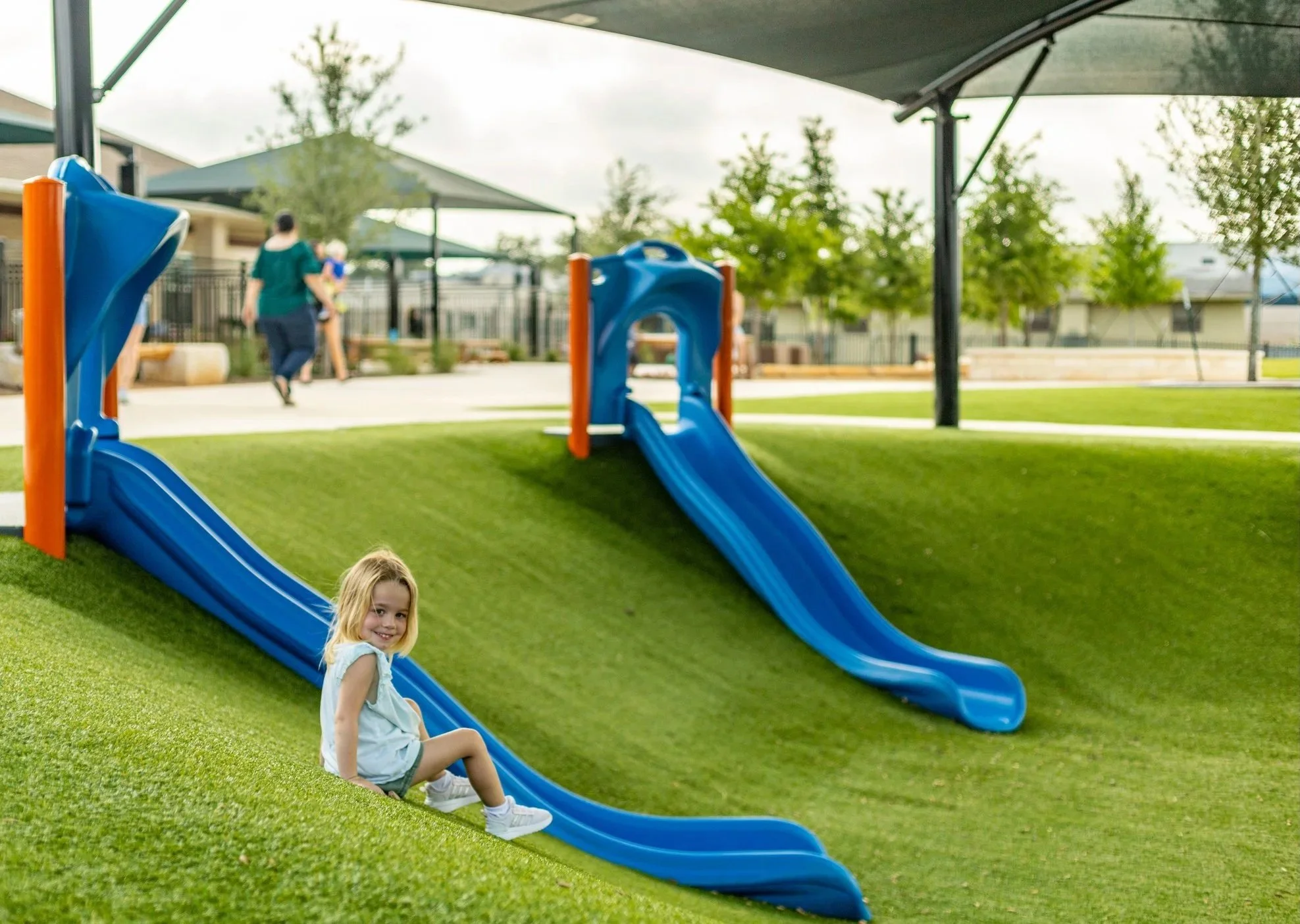 Young girl sitting on a small blue slide at a playground with artificial grass, in a park with trees and shaded areas, and a woman walking with a child in the background.