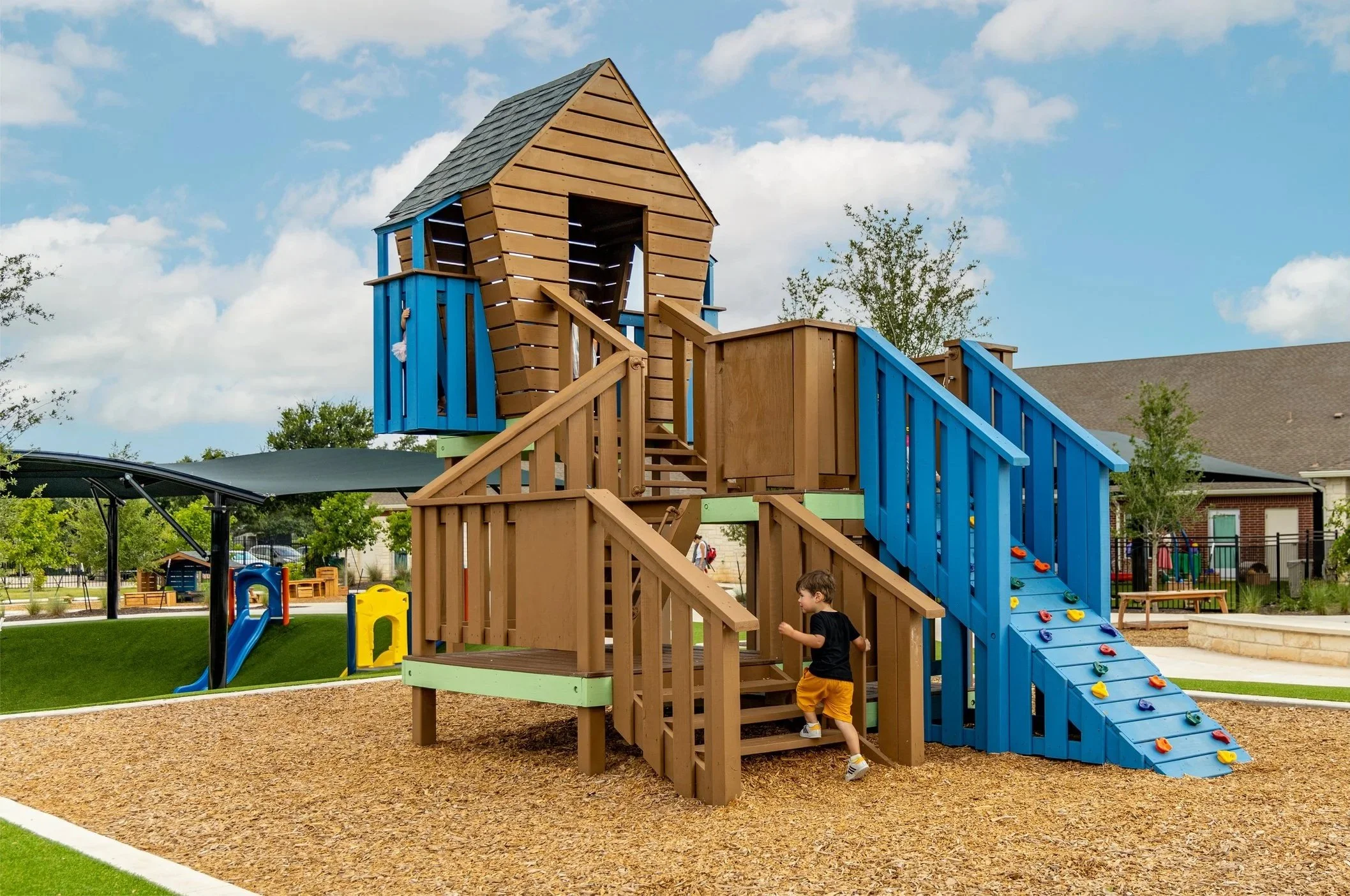 Children's playground with a multi-level wooden play structure featuring a climbing wall, slide, and lookout tower on a gravel surface, surrounded by grassy areas and trees.