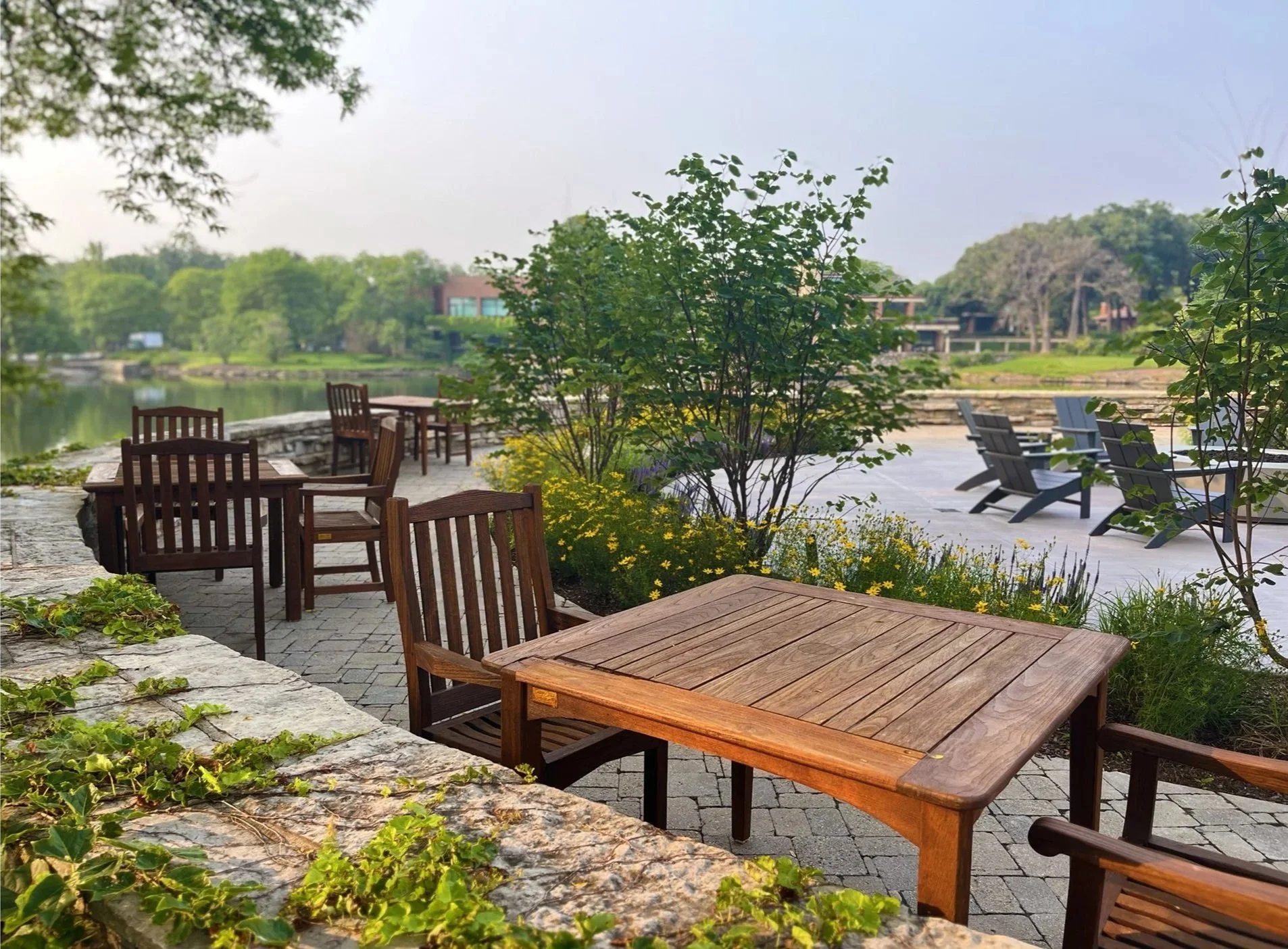 Outdoor seating area with wooden tables and chairs near a lake, surrounded by green trees and shrubs, with Adirondack chairs on a patio in the background.