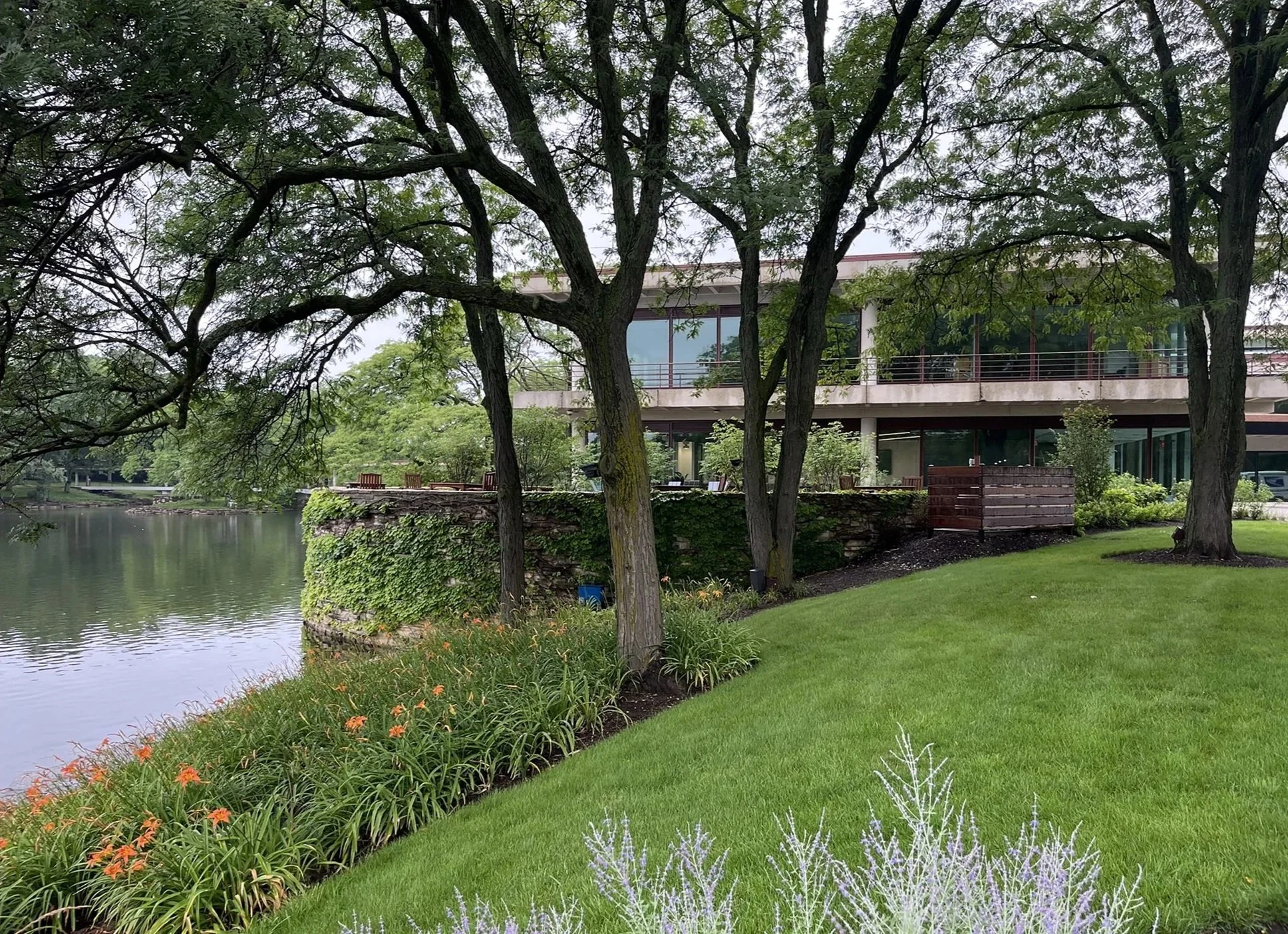 Modern glass building overlooking a river, surrounded by lush green trees, grass, and landscaped flowers, with an outdoor patio area.