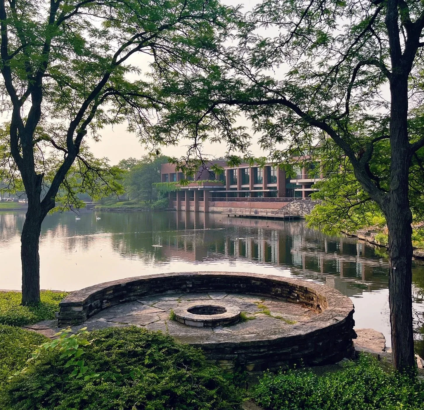 A landscape featuring a calm river with a building on the far side, two trees framing the scene, and a stone fire pit with seating area in the foreground.
