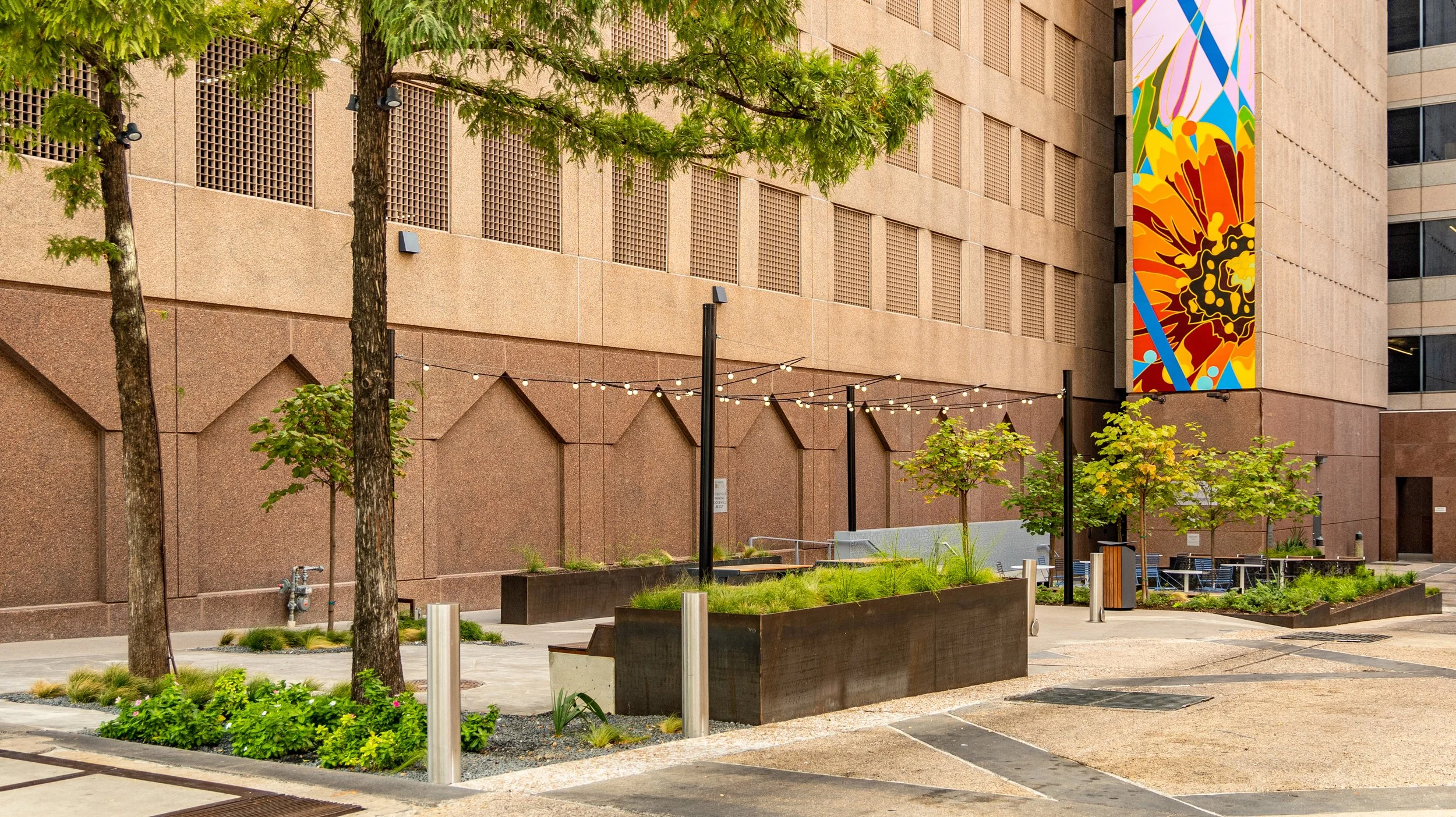 Urban outdoor courtyard with trees, plants, benches, string lights, and a colorful mural on the building wall.