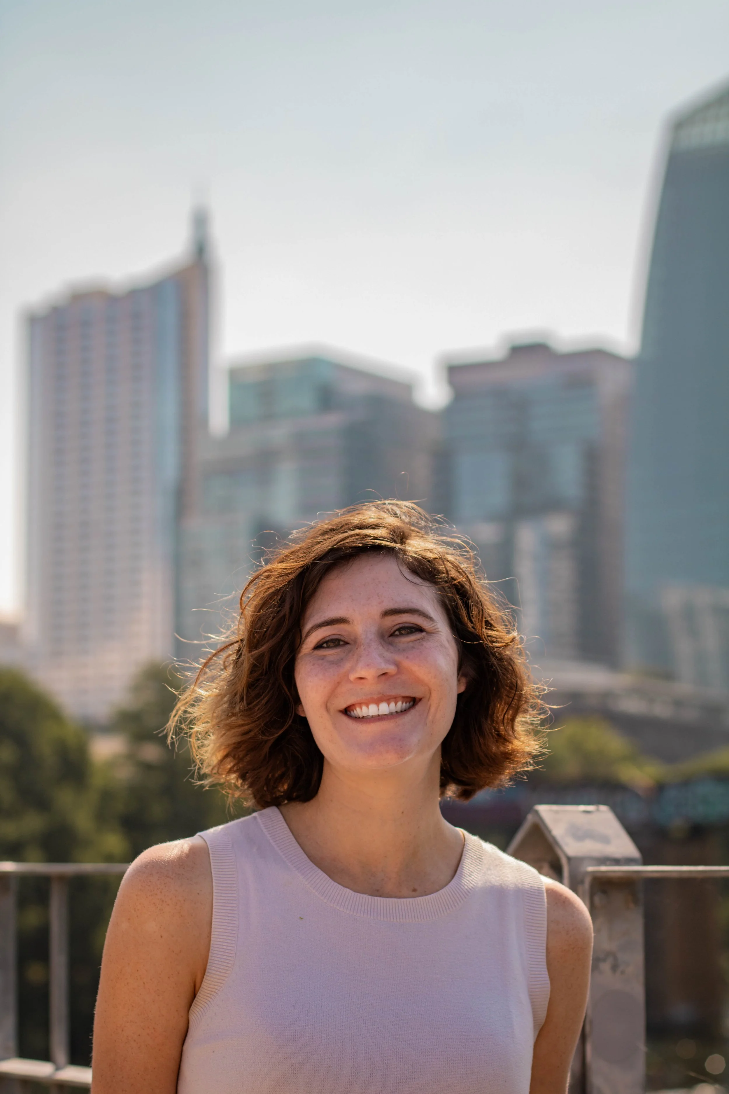 A smiling woman with short curly brown hair and freckles, wearing a sleeveless beige top, standing outdoors with city buildings and trees in the background.